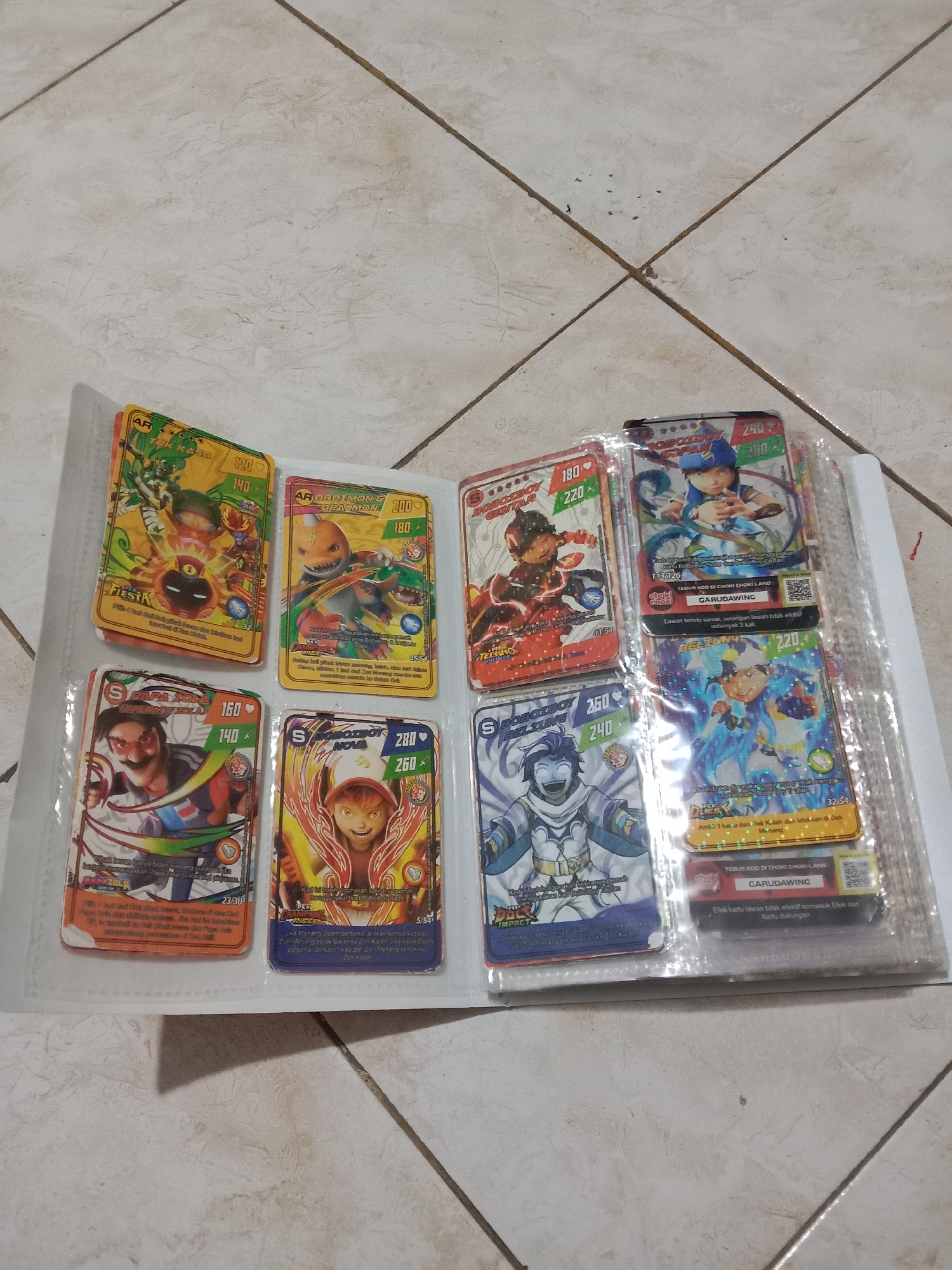 The image shows an open binder with trading cards. The binder is on a tiled floor with a light beige color and diagonal grout lines. The cards are colorful, featuring animated characters and text in Indonesian. Some of the visible text includes "AR OBOKIMON G OPALMON," "S PAPA ZOLA," "S BOBOTBOT MELTUNG," and "BOBOTBOT CENTUR." The scene is indoors, with no visible indication of the city of Madiun, Indonesia.
