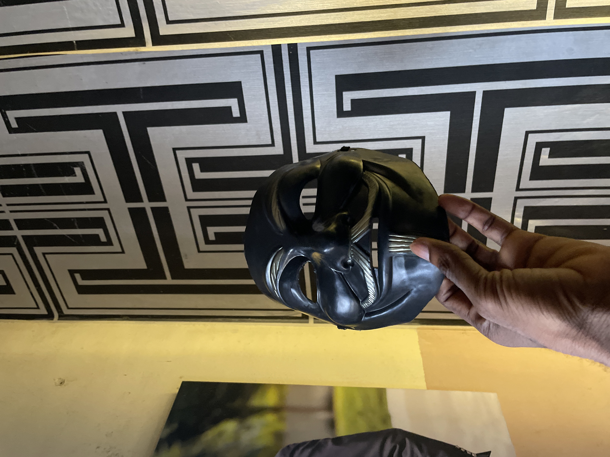 A black mask with silver accents is held in a person's hand against a backdrop of geometric patterned wallpaper. The wallpaper features silver and black rectangular designs. The scene is lit with artificial light, creating a yellowish cast on a wall below the wallpaper. A corner of a framed photograph or artwork is partially visible below the mask and the wall. The location is unknown.