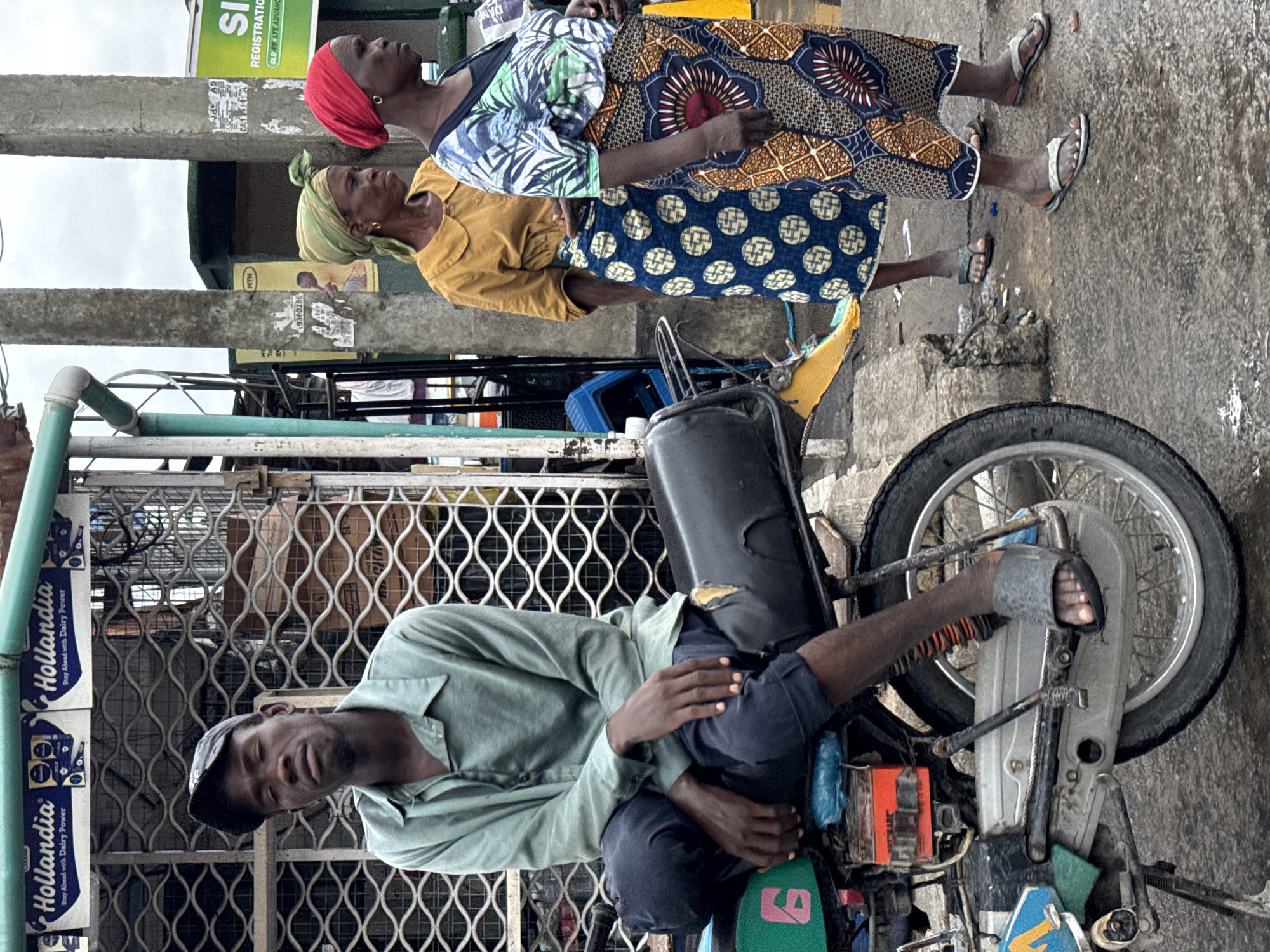 The image captures a street scene in Ado, Nigeria. A man reclines on a motorcycle, resting his head near a closed shop front protected by a metal gate. Boxes of "Hollandia" dairy products are stacked along the gate. Two women stand on the wet street, wearing traditional clothing and headscarves. A sign above them advertises "SI" registration and GLO 4G LTE. The location appears urban and bustling.