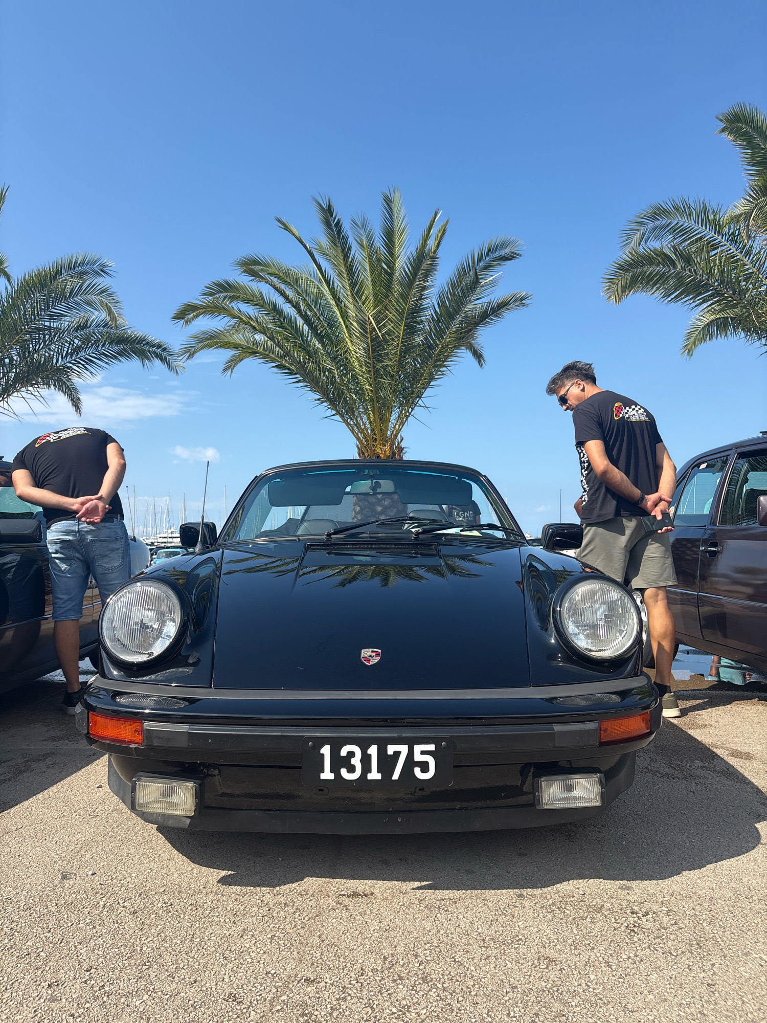 In Bar, Montenegro, a black Porsche 911 convertible is parked outdoors under a clear blue sky, with palm trees visible in the background. Two men are standing near the car. The man on the left, wearing a black t-shirt and denim shorts, is facing away from the camera, looking at the car. The man on the right, also in a black t-shirt and khaki shorts, is leaning over and inspecting the front of the Porsche. The car's license plate reads "13175". The ground is covered in asphalt or gravel. Boats are visible docked in the background, suggesting the car is parked near a harbor or marina.