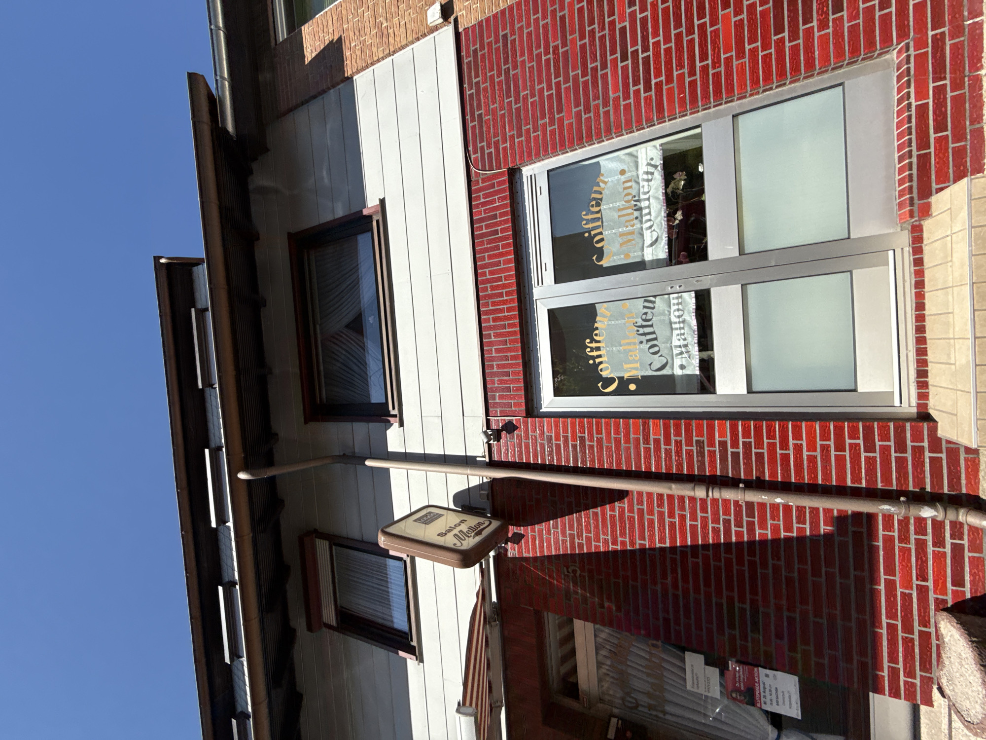 A low-angle, wide shot shows the exterior of a two-story building with a red brick lower facade and white horizontal siding on the upper story. The building is situated under a clear blue sky. A brown downspout runs vertically along the brick wall. A sign hanging from a pole, reads "Salon Mallon Coiffeur". Two large glass doors are visible on the ground floor, with text that appears to say "Coiffeur Mallon Coiffeur". Above the brickwork, on the upper white siding, are two windows with dark frames. The building has a modern architectural style with visible rooflines and eaves.