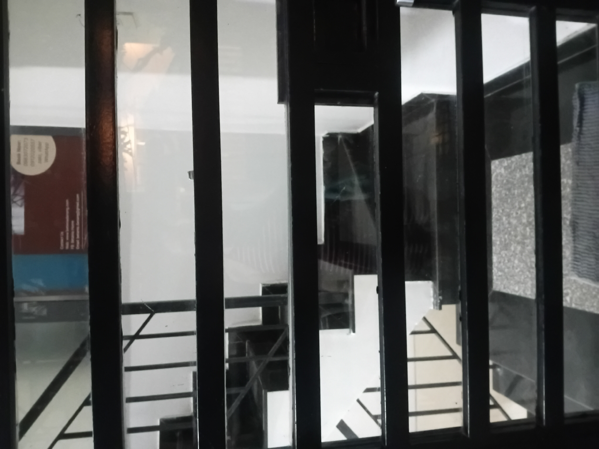 The media file shows a view through a dark metal grate, looking into an interior space. The grate consists of several vertical bars and horizontal elements, creating a series of rectangular openings.
Inside, the scene appears to be a modern building with a staircase. The staircase is visible in multiple sections through the grate openings. It has white steps and dark railings. Some parts of the staircase are well-lit, while others are in shadow. The walls appear to be white. In one of the openings on the left, there is a glimpse of a dark object with some text on it, possibly a poster or signage, with a circular white label on the top left. Further into the building, dark panels or furniture are visible. The overall impression is one of an indoor staircase in a contemporary setting.
The time of day is not discernible, nor is the weather. The location context suggests Đà Nẵng, Vietnam, but there are no specific visual cues within the image to confirm this. The scene is static, with no discernible activity or people. The lighting suggests artificial illumination inside the building. The mood is neutral, with the grate obscuring a full view and creating a sense of separation or observation.