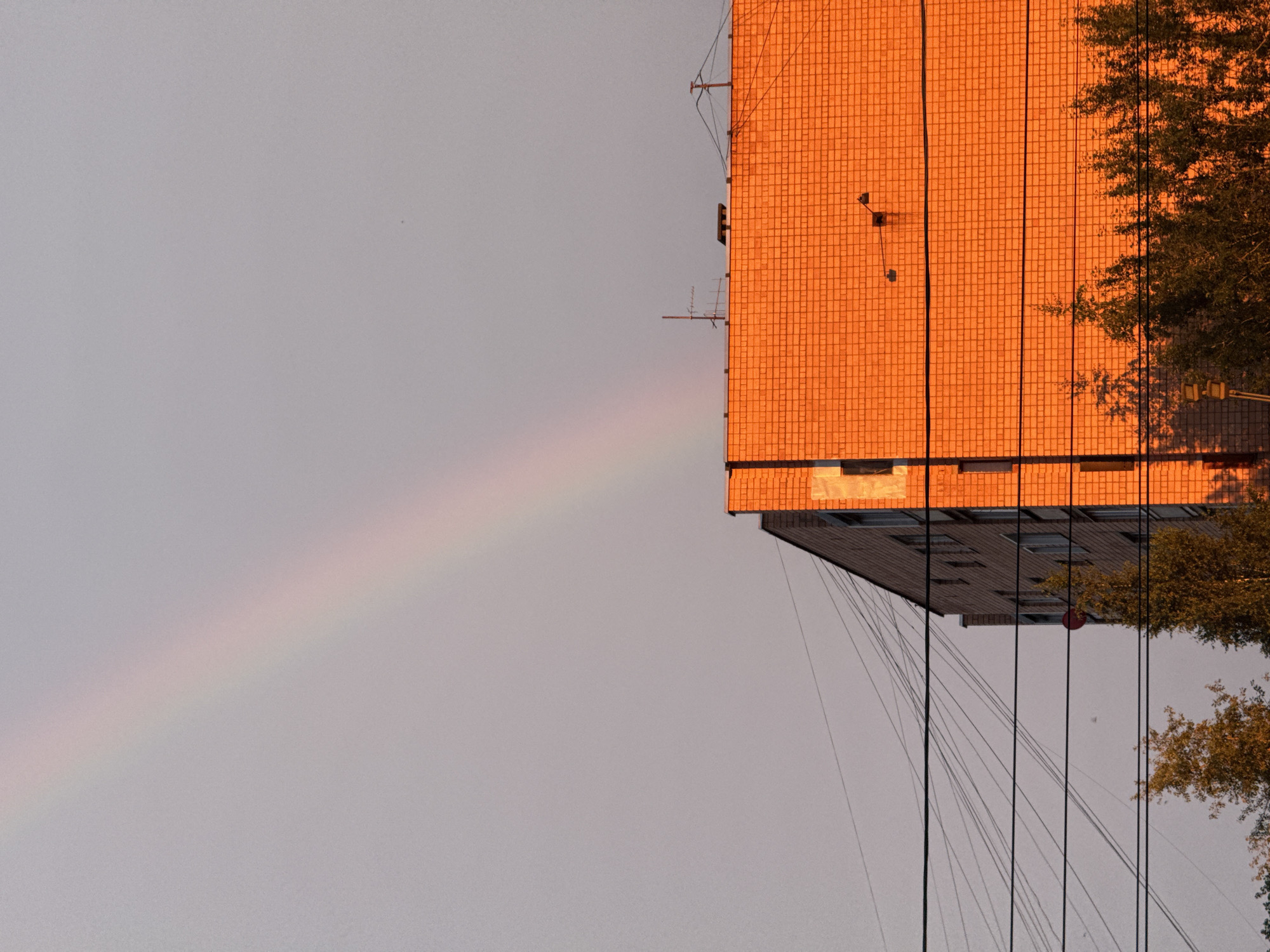 This image depicts a building with an orange tiled facade, illuminated by the warm glow of the setting sun. A faint rainbow arcs across the pale sky to the left of the building, suggesting recent rain. The perspective is from below, looking up at the structure and the sky. Electrical wires hang down from the building, creating a network of lines against the sky and the building's lower levels. A tree with green foliage is visible on the right side of the frame, casting a shadow. The building appears to be residential, with visible windows on its lower section. The scene is quiet and still, with a serene atmosphere. The time of day is likely late afternoon or early evening, given the golden light and the presence of a rainbow. There is no visible text in the image.