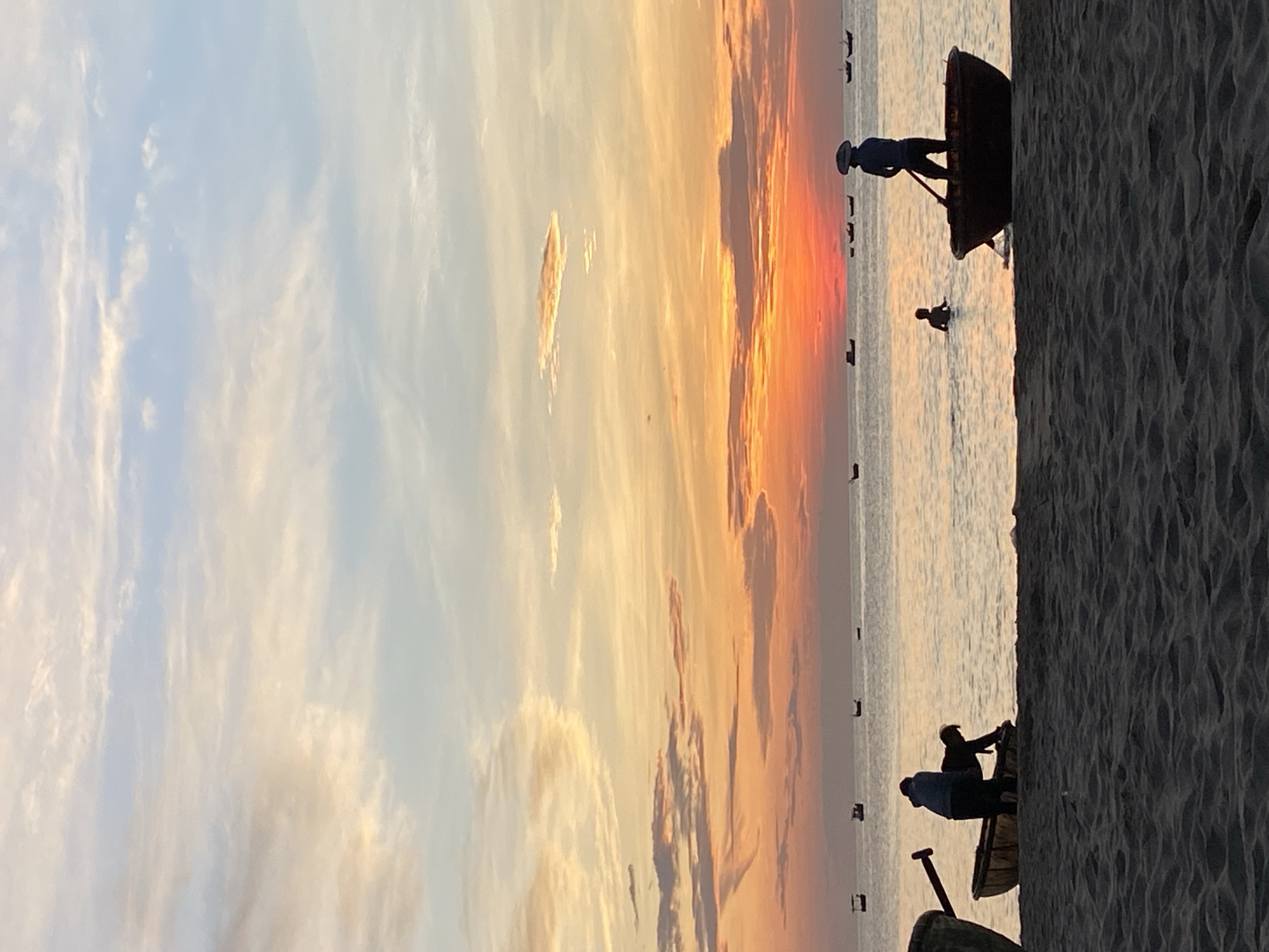 At sunset on a beach in Da Nang, Vietnam, several people are in and around traditional basket boats. The sky is a vibrant mix of orange, pink, and blue, with scattered clouds. In the foreground, a sandy beach stretches across the bottom of the frame. Closer to the water, two individuals are bent over a basket boat, appearing to be working on it or preparing it for use. Further out in the calm sea, a person stands in a basket boat, holding a paddle, and another person is wading in the shallow water. Numerous other small boats are scattered across the horizon. The overall atmosphere is peaceful and serene, capturing a typical evening scene on the Vietnamese coast.