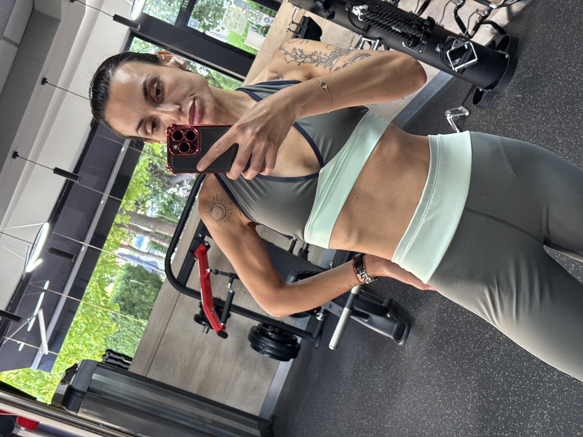 A young adult woman is in the interior of a gym. She is holding a smartphone with a red case and is taking a selfie. She wears athletic wear, including a sports bra with light green trim and leggings. Tattoos are visible on her arms. Gym equipment surrounds her, including a weight machine and a treadmill. Outside the windows, trees are visible. The location may be the city of Pleven, Bulgaria.