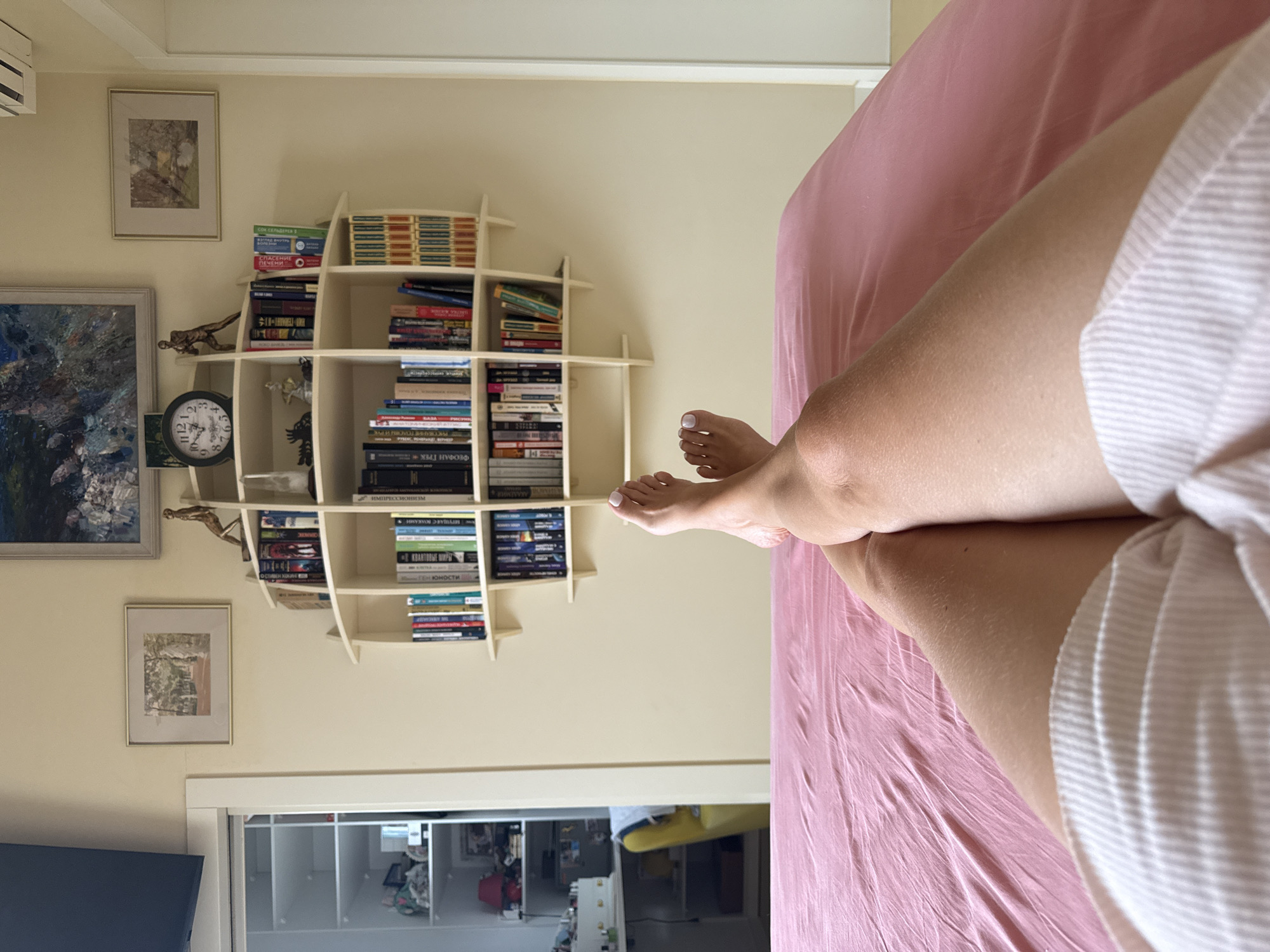 A first-person perspective shot from a bed shows crossed legs with white-painted toenails lying on a pink sheet. In the background, a wall is decorated with two framed landscape paintings and a round analog clock. A white, multi-tiered, curved bookshelf is mounted on the wall, filled with numerous books. To the left of the bookshelf, a glimpse of a white closet with glass doors reveals items inside. The overall scene suggests a bedroom interior. The mention of Zimyonki (Zimenki, Zimenka), Russia, is not supported by any visual evidence in the image.