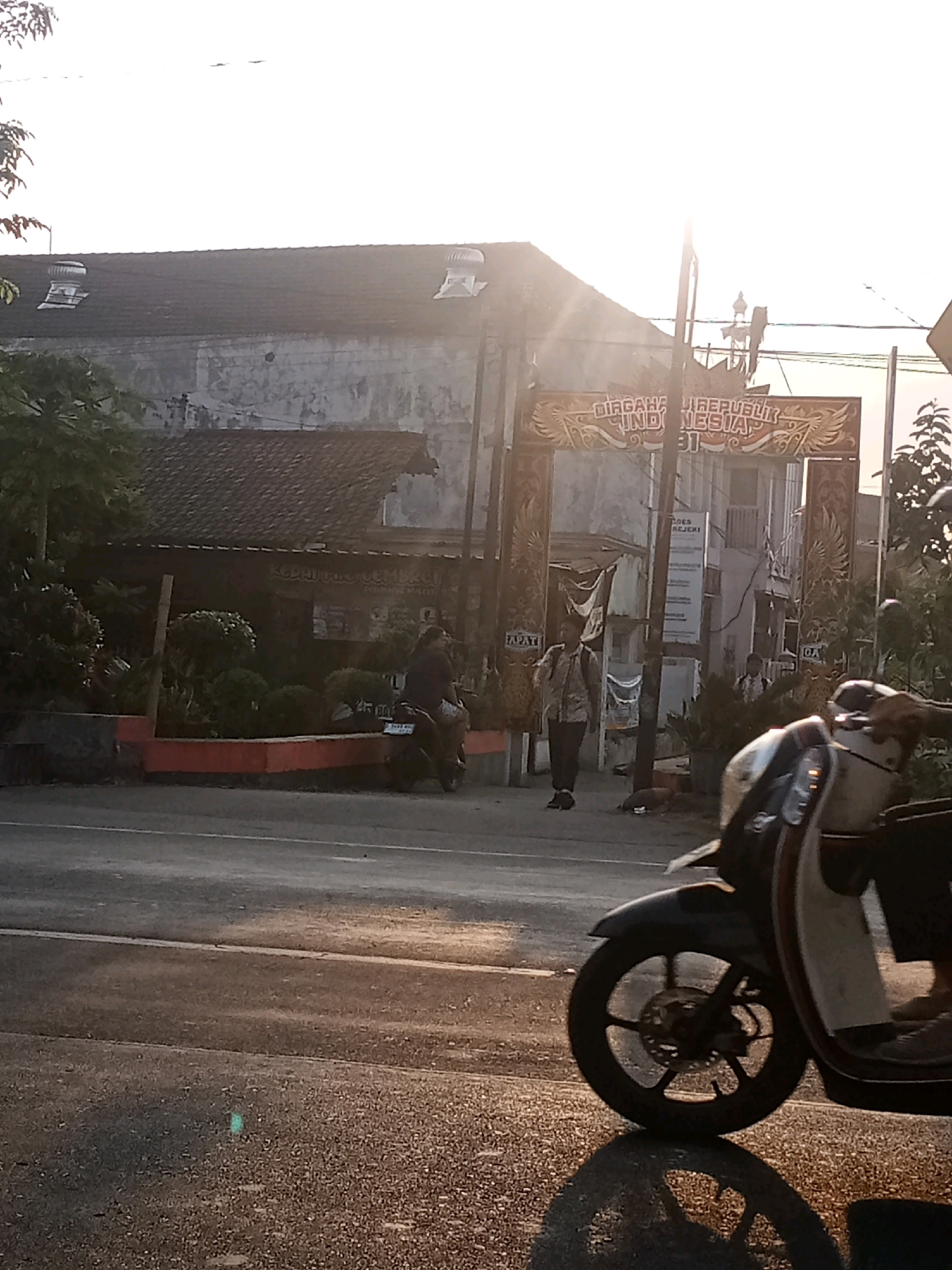 The image depicts a street scene in Kaliwungu, Indonesia, likely during the late afternoon or early evening, given the low angle of the sun and the long shadows. In the foreground, a dark-colored motorcycle is partially visible, with its front wheel and a portion of its body in focus. The motorcycle is parked on the right side of the frame, casting a reflection on the wet asphalt of the road.
Behind the motorcycle, a two-lane road stretches across the image, with a white line marking the lanes. The road surface appears wet, possibly from recent rain, and reflects the ambient light.
In the midground, an arched gateway stands prominently. The gateway is decorated with ornate patterns and text in Indonesian. The text on the gateway reads "DIRGAHAYU REPUBLIK INDONESIA 71," which translates to "Long Live the Republic of Indonesia 71," indicating a celebration of Indonesia's Independence Day. Two individuals are visible near the gateway. One person, wearing a patterned shirt and dark pants, stands on the left side of the gateway, facing towards the street. Another person, wearing a light-colored shirt and dark pants, is seen further back, walking under the gateway. A vertical signpost with text is also present near the gateway.
To the left of the gateway, a building with a tiled roof and weathered walls is visible. Some vegetation, including trees and bushes, is planted along the front of this building, with a red-painted border. A person is riding a motorcycle on the road, heading away from the viewer and towards the gateway.
In the background, more buildings and trees are visible, with the sky a bright white, suggesting the sun is low in the sky or that the image is overexposed in that area. The overall atmosphere is one of a typical Indonesian street scene, possibly on a national holiday.
