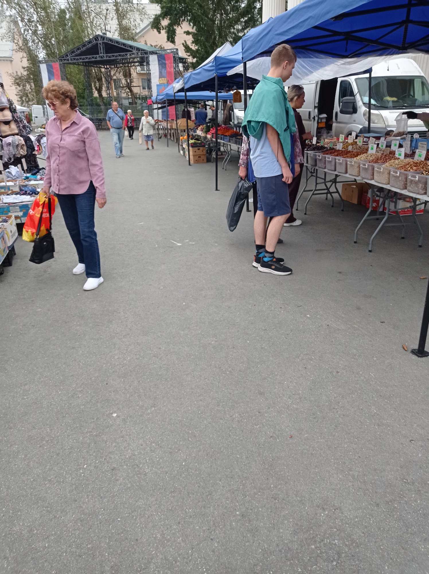 The image depicts an outdoor market scene in Degtyarsk, Russia. In the foreground, a woman with curly brown hair and glasses, wearing a pink shirt and blue jeans, walks while holding a red and yellow shopping bag and a black purse. To the right, a young man in a teal overshirt, blue t-shirt, and navy shorts stands near a market stall displaying various nuts and dried fruits. Several other individuals are visible in the background, along with market tents, a white van, and a stage structure adorned with Russian flags. The ground is paved asphalt.