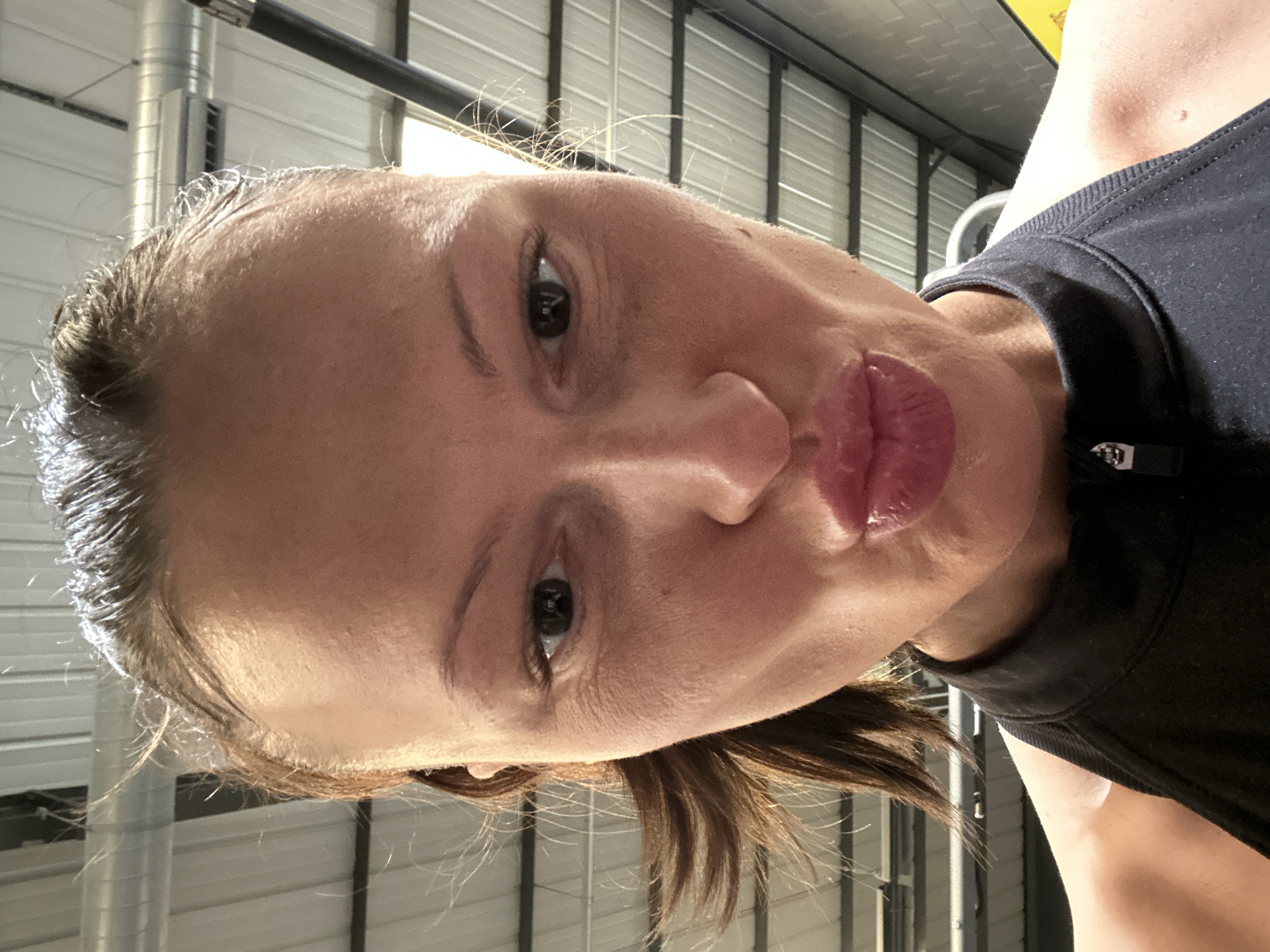 A close-up, eye-level photographic image depicts a female subject positioned centrally, directly facing the camera. The subject has light brown hair, pulled back, with individual strands visible around the hairline. Her facial skin exhibits a moist sheen. Her lips are distinctly pursed, forming a pouting expression, while her eyes are open and directed at the lens. She wears a black, sleeveless athletic garment featuring a zippered high collar. The background is rendered out of focus, presenting an interior setting with a high, corrugated metal ceiling. Silver-colored cylindrical ventilation ductwork is visible horizontally across the ceiling area. A dark, vertical structural element is partially discernible on the right side of the frame, and a portion of a yellow object is visible in the lower right background. This environment is characteristic of an indoor fitness facility, specifically a gymnasium, located within the municipal area of Tonbridge, within the Tonbridge and Malling borough, United Kingdom.