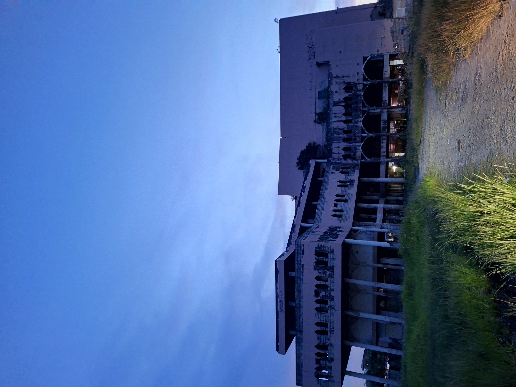 The image shows an exterior view of a large, multi-story, unfinished pink concrete building in Lhokseumawe, Indonesia. The building has arched openings and columns along its lower levels. The sky is a gradient of blue hues. A dirt path and grassy area borders the building.
