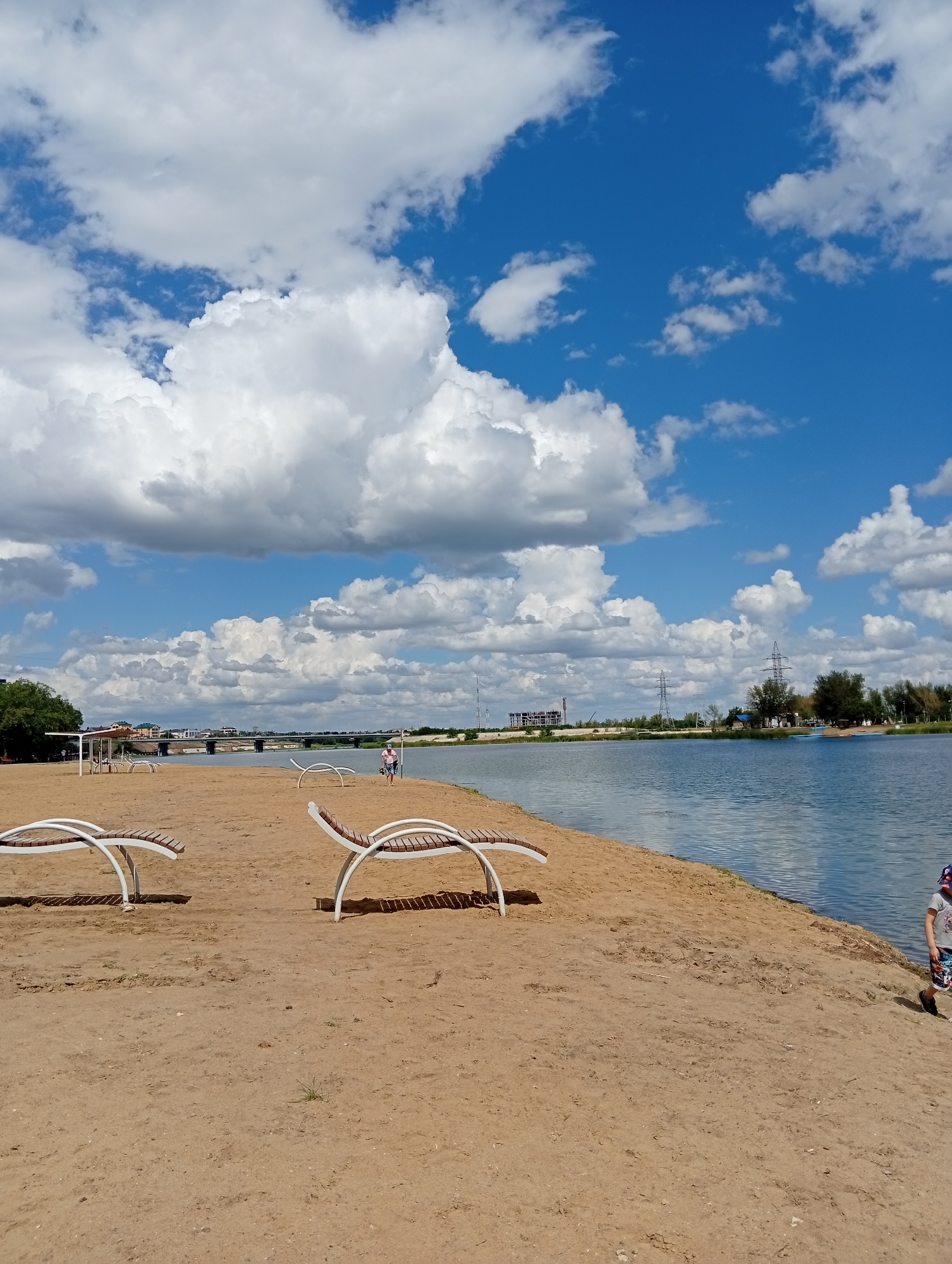 A wide-angle, eye-level shot captures a sandy beach along a body of water under a bright, cloudy sky. In the foreground, two empty white and brown lounge chairs sit on the sand. To the right, a young person wearing a hat, t-shirt, and shorts walks away from the camera towards the water. The water's surface is calm and reflects the sky. In the midground, a person is walking on the beach near a few other lounge chairs and a small shelter. Across the water, a bridge spans the distance, with buildings and industrial structures visible on the far bank. The sky is predominantly blue with large, fluffy white clouds. The scene is set in the Zarechnyy Rural District, Kazakhstan.