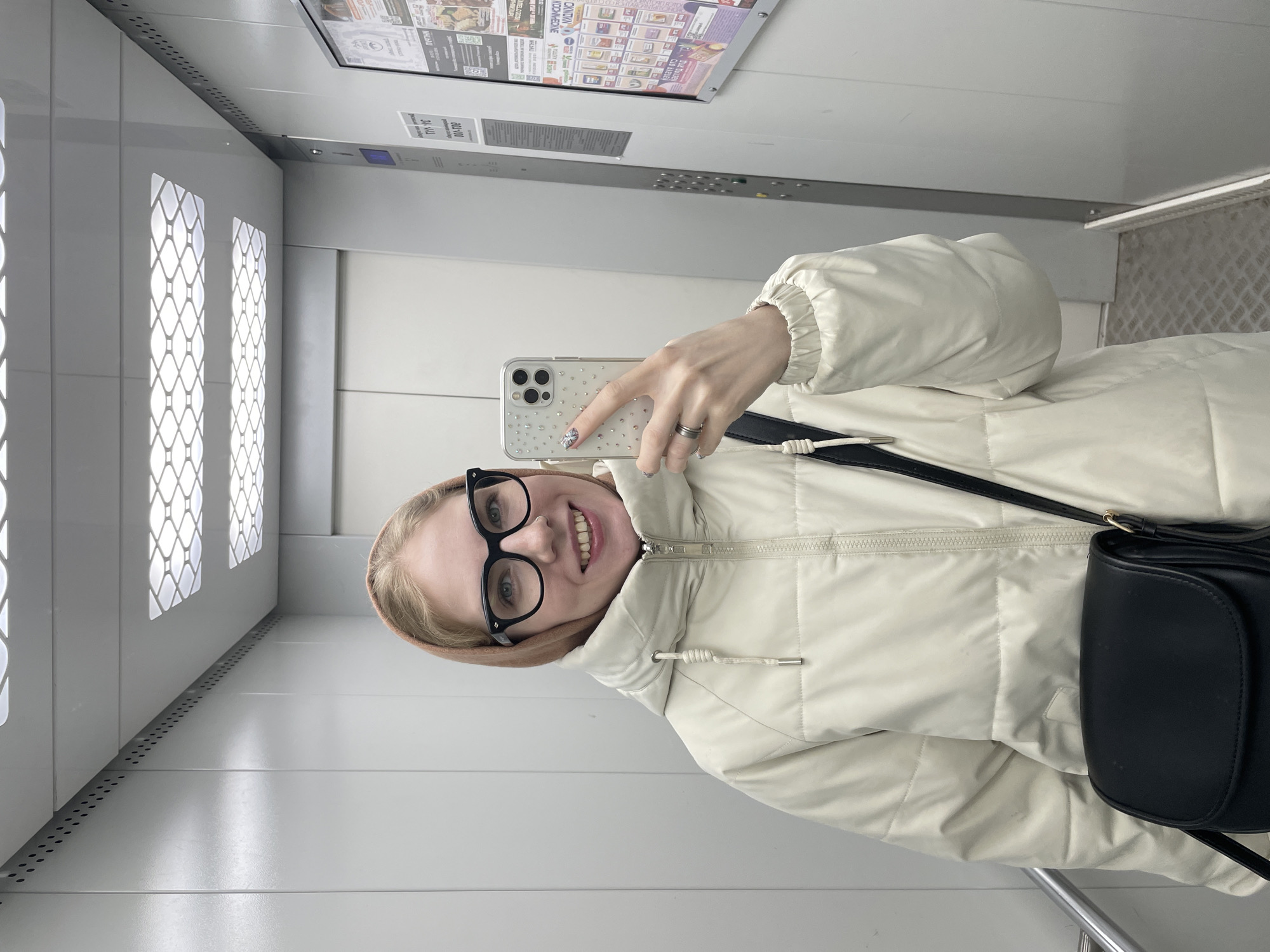 A young woman is taking a selfie in what appears to be an elevator. The elevator has a modern interior with white walls, decorative panels, and a control panel. The woman is wearing glasses and a light-colored, puffy jacket. She has a black purse slung across her body. A poster or informational display is visible on the elevator wall behind her. Given the request for location and the user history, the image is assumed to have been taken in Izhevsk, Russia. The elevator environment seems clean and well-maintained.
