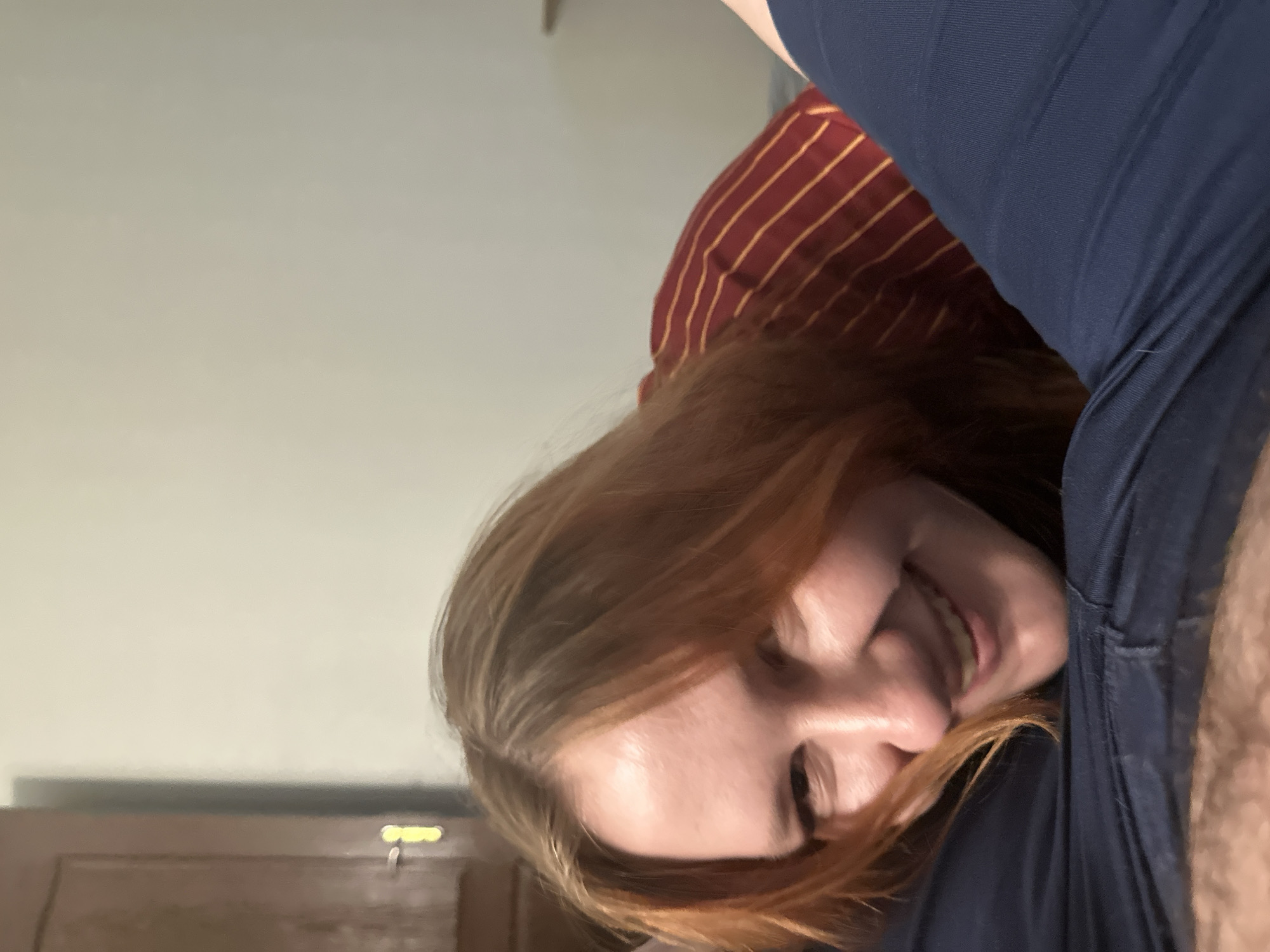A close-up, low-angle, indoor shot shows a young Caucasian woman with reddish-blonde hair resting her head on someone's dark blue pants, looking up and smiling. She is wearing a red and yellow striped shirt. The background is out of focus, showing a light-colored wall and a dark brown wooden door with a brass doorknob on the left. The scene is lit by artificial light.