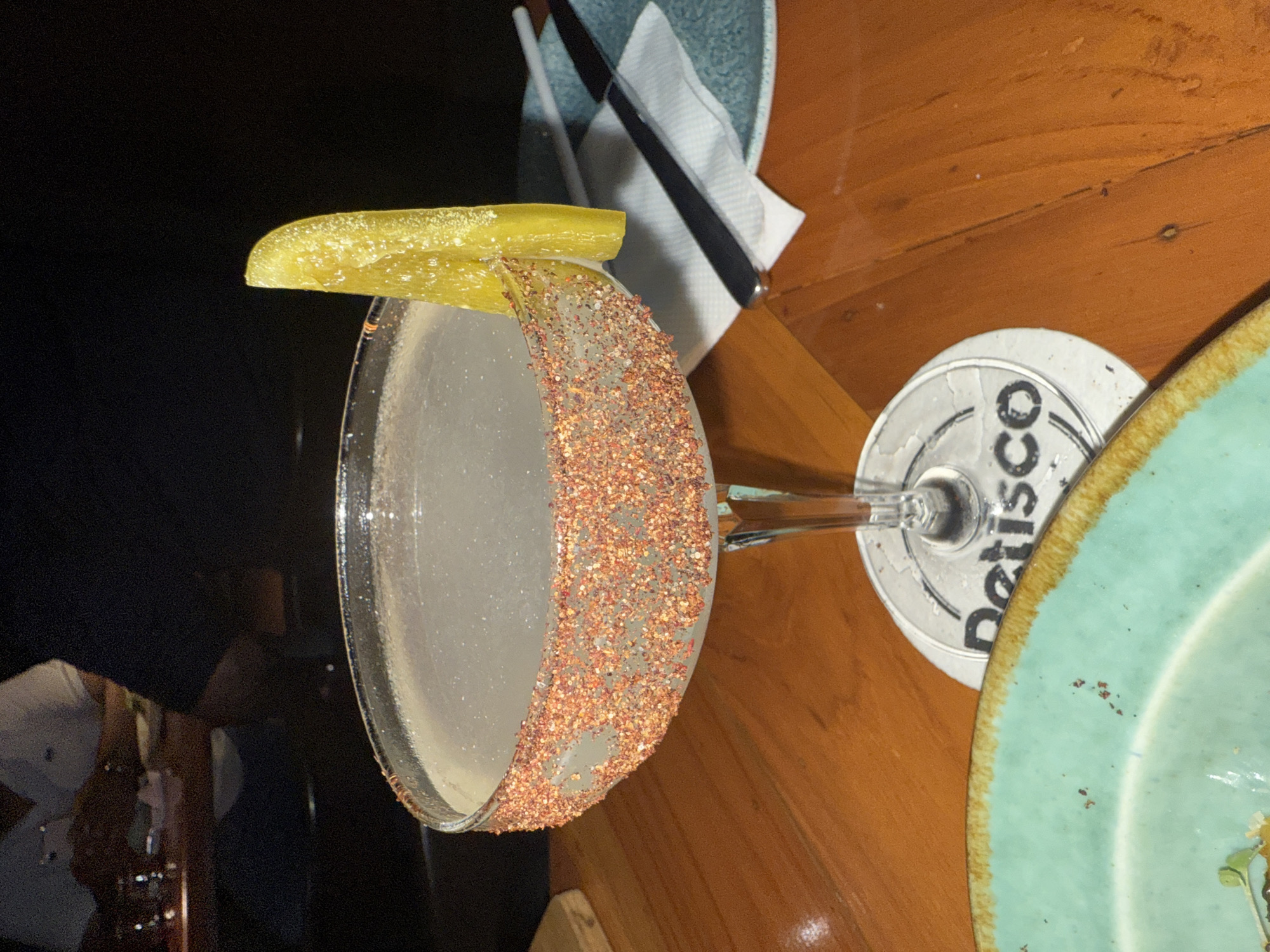 Here is a description of the image:
The image shows a cocktail in a coupe glass with a chili-lime salt rim and a pickle garnish on a wooden table. A coaster with the word "Detisco" is visible under the stem of the glass. Part of a plate with a turquoise rim is also visible. Utensils wrapped in a napkin are in the background. Based on the coaster and general aesthetic, it is possible that the scene is located in Panaji, India.