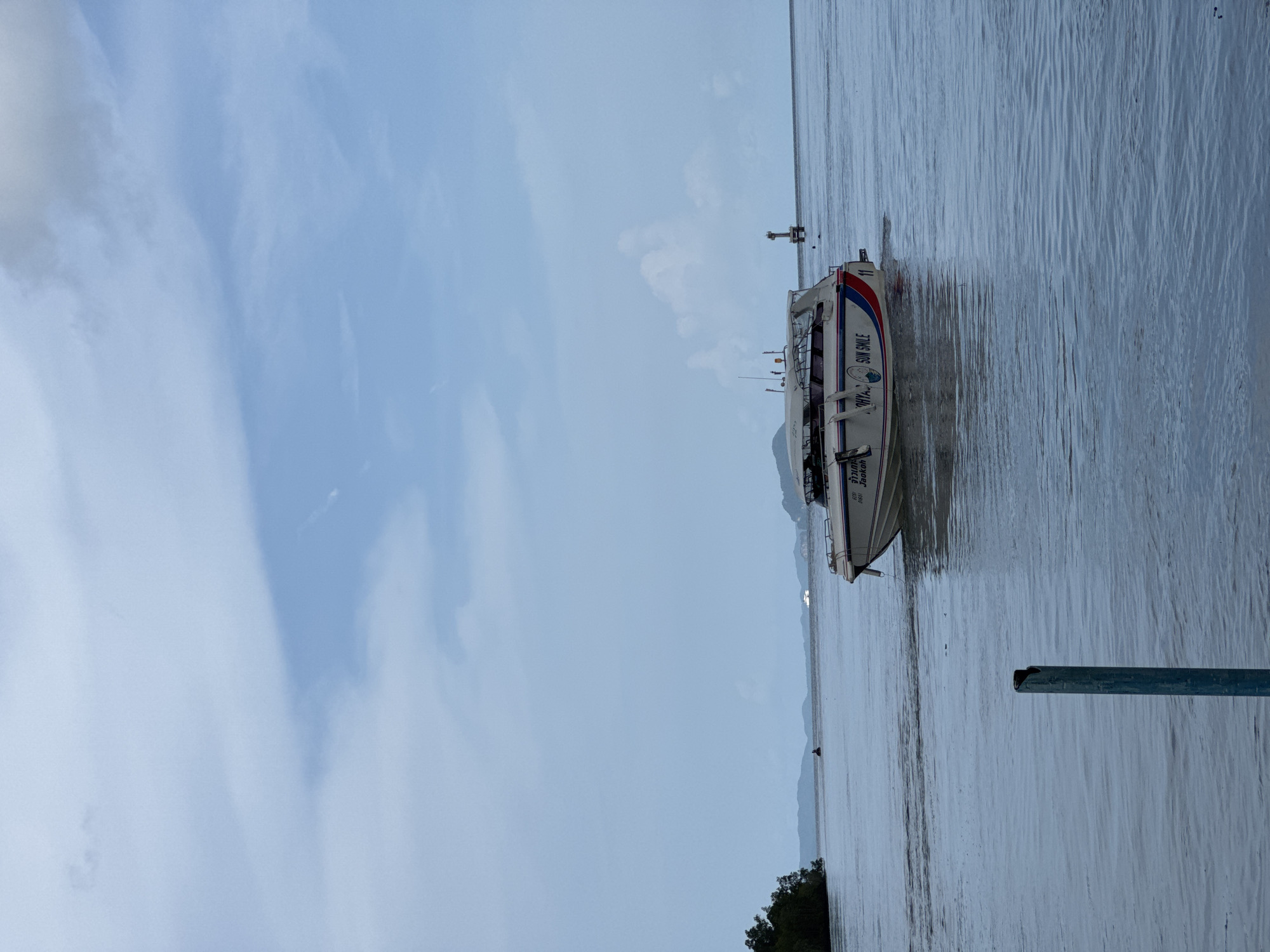 A speed boat labeled "SUN SMILE" travels across calm, grey water under an overcast sky. The boat, decorated with red and blue stripes, has a white cabin with dark-tinted windows. In the distance, islands with green foliage are visible on the horizon. A lighthouse with a white structure on a dark base stands in the water to the right of the boat. In the foreground, a faded blue pole rises from the water. The scene appears to be the coast of Pa Khlok, Thailand.