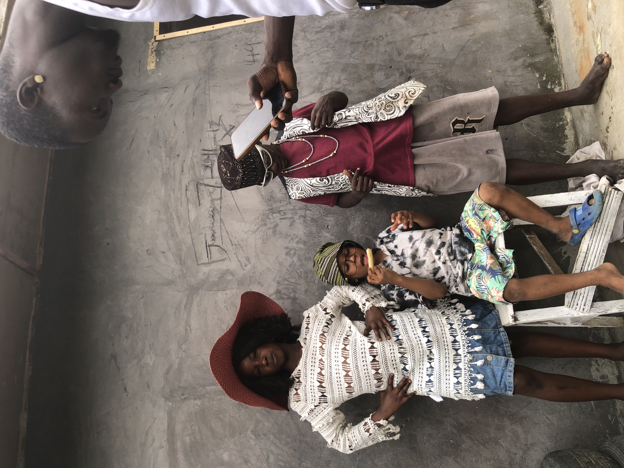 In Ado, Nigeria, a group of four individuals is captured in a candid outdoor scene against a textured concrete wall. In the foreground on the right, a man, partially visible, holds a smartphone, appearing to film or photograph the scene. He wears a white tank top. To his left, a young person with dark skin is styled in a burgundy t-shirt, a patterned vest, light brown shorts with a prominent "R" logo, a tall, patterned hat, and white-framed sunglasses. They are adorned with two beaded necklaces. To the left of this individual, a young person with dark skin wears a white, open-weave long-sleeved top over a denim skirt and a wide-brimmed red hat. Next to them, another young person, also with dark skin, is seated on a white step stool. They are wearing a tie-dye t-shirt, colorful patterned shorts, and a green and yellow striped hat. This individual is holding a piece of food to their mouth. The environment includes the rough concrete wall with faint markings and graffiti, and the ground is a mix of concrete and dirt. The overall atmosphere suggests a casual, possibly artistic or performance-related gathering.