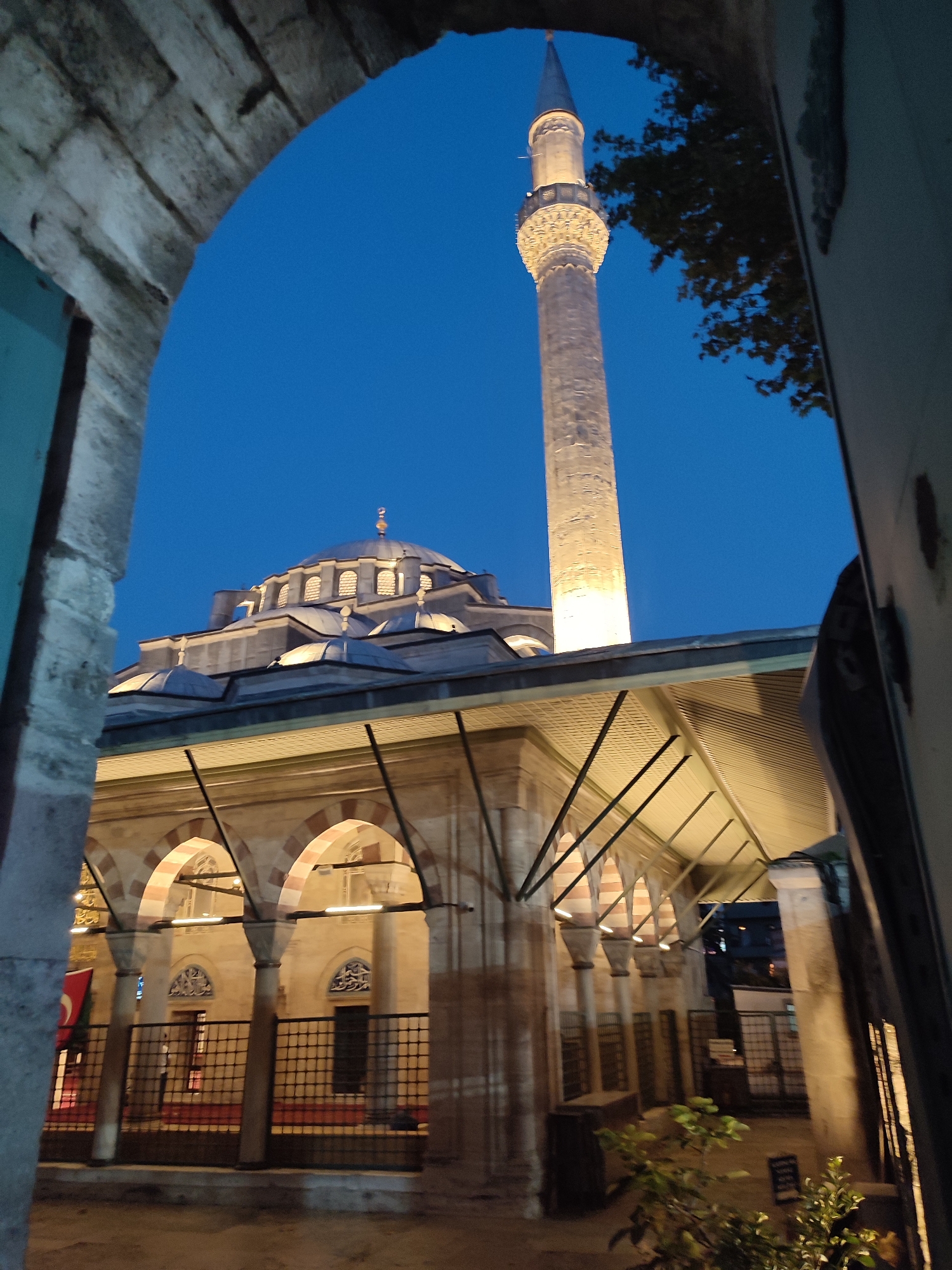 The image depicts a view of a mosque in Beyoğlu, Turkey, framed by an arched entryway. The mosque features a prominent minaret illuminated by warm light against a deep blue twilight sky. The main building has a large dome and several smaller domes, all intricately designed and lit. Below the domes, the mosque's facade is characterized by a series of arches supported by pillars, with patterned stonework. Through the arched openings, a courtyard with red carpeting and intricate window designs is visible. A Turkish flag hangs on the left side of the courtyard. The scene is captured from a slightly elevated perspective, looking towards the mosque.