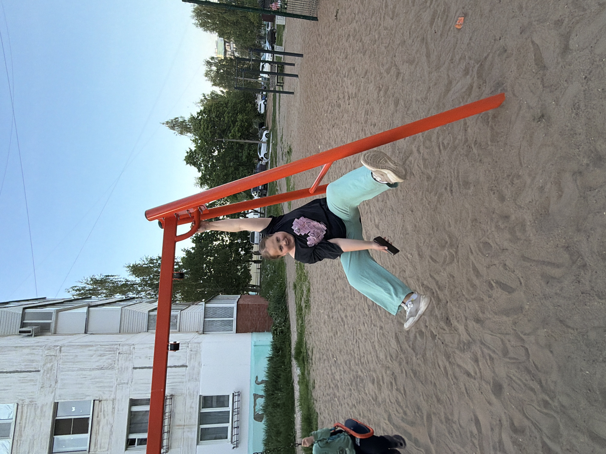 A woman with a smartphone in her hand is suspended upside down on a red metal playground structure in what appears to be a sand pit. She is wearing a black t-shirt with a design on it, light green pants, and gray sneakers. The playground is located in a city, evident from the apartment building visible in the background with white walls and windows. Trees and parked cars are also visible, suggesting a park or residential area. The image was taken in Chaikovsky, Russia, as evidenced by the architectural style of the building and the presence of Cyrillic text on the building itself.