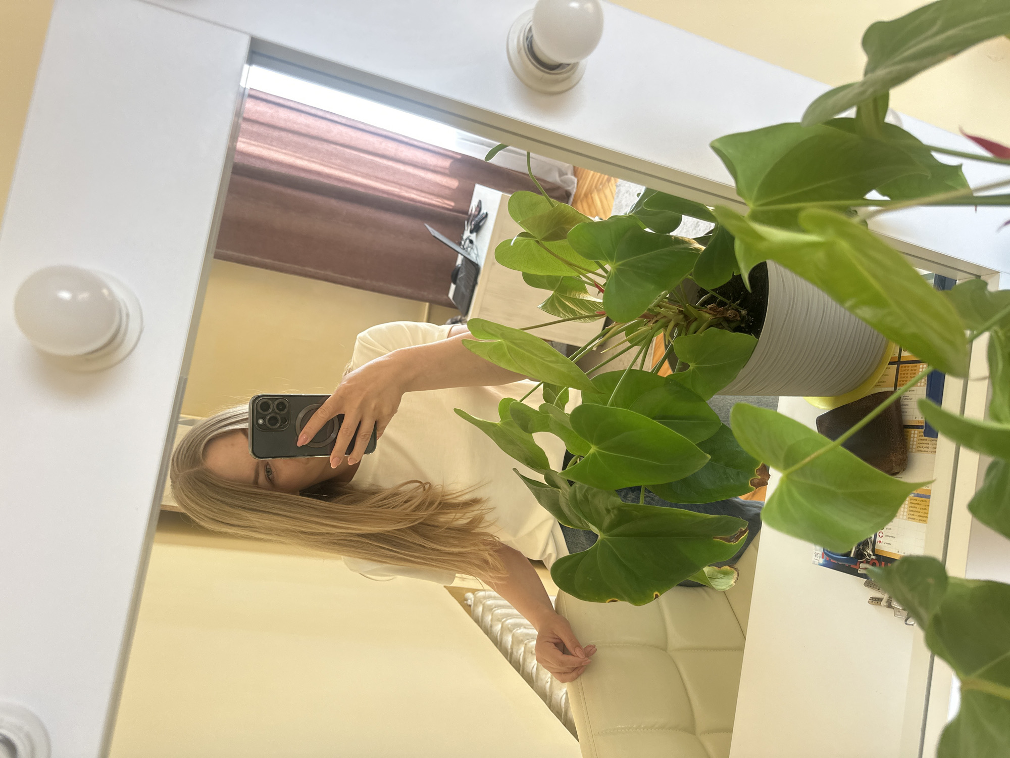 A young woman with blonde hair takes a mirror selfie in a room with light yellow walls and a light brown curtain. She is wearing a white t-shirt and blue jeans and is holding her phone in her right hand. In front of her, on a white surface, is a potted plant with large green leaves. The room appears to be a bedroom or dressing area, with a makeup mirror and lights visible in the reflection. The scene is indoors, and there is no indication of the specific location within Ternopil, Ukraine.