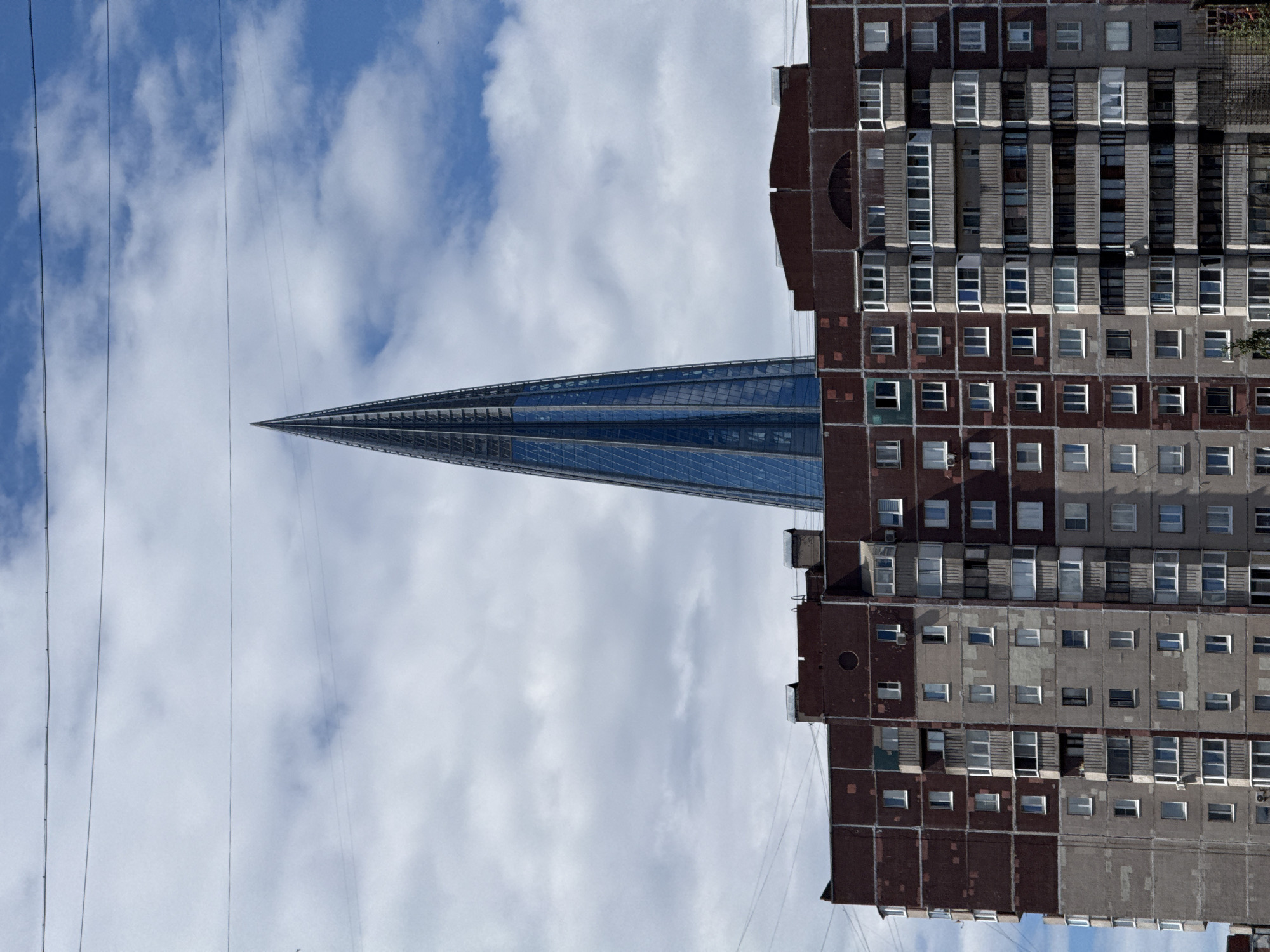 This vertical, eye-level shot captures a juxtaposition of modern and older architecture against a cloudy sky in Saint Petersburg, Russia. The primary subjects are two buildings: a sleek, pointed skyscraper with a glass facade, and a weathered, multi-story brick apartment building with numerous windows. The sky is a mix of blue and white clouds, suggesting daytime, possibly late morning or early afternoon.
The modern skyscraper, with its sharp, angular design, dominates the left side of the frame, its glass surfaces reflecting the sky and clouds. It tapers to a needle-like point at the top. Adjacent to it, on the right, is the older apartment building, characterized by its reddish-brown brick and concrete facade, showing signs of age and wear. The apartment building is filled with rows of windows, some of which appear to be air conditioning units installed. Several power lines crisscross the scene, adding a utilitarian element to the urban landscape.
There is no discernible activity or event taking place; the image appears to be a static depiction of the city's architecture. The weather is overcast but bright, typical of Saint Petersburg. The contrast between the futuristic design of the skyscraper and the more utilitarian, somewhat aged appearance of the apartment building is a notable detail, highlighting the city's evolving urban fabric. No text is visible in the image.