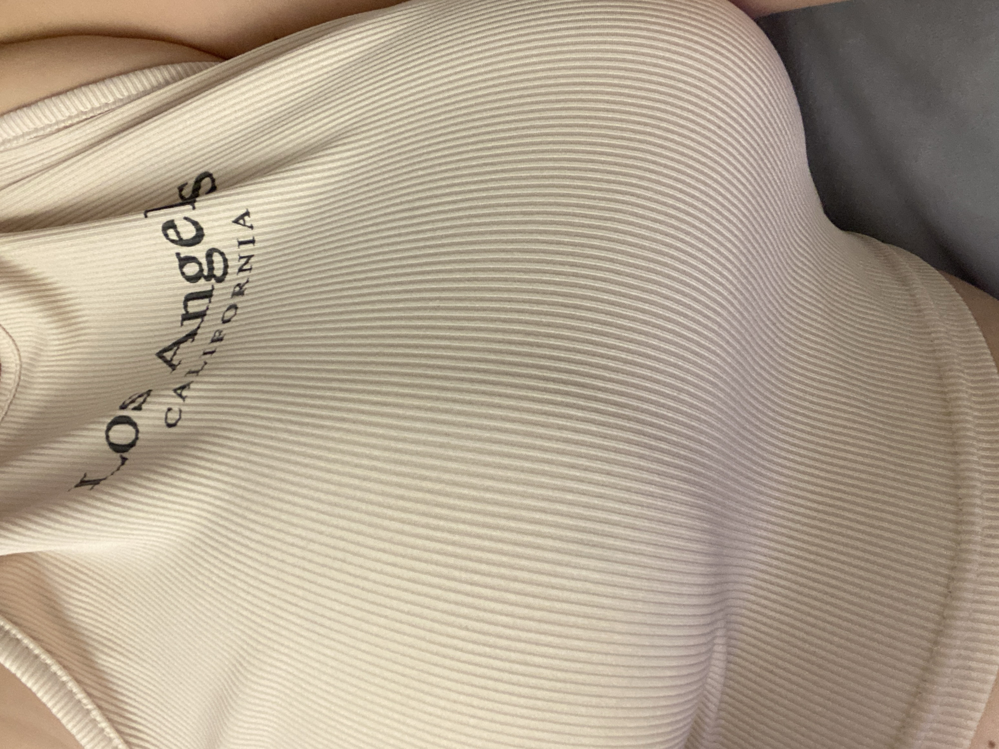 The image shows a close-up of a beige ribbed tank top with black text. The text reads "Los Angeles California". The tank top appears to be worn by a person, with a portion of their skin visible on the right side of the image. The background is a dark, blurry surface, possibly a piece of furniture. There is no visible indication of the city of Perm, Russia, or any specific location within it.