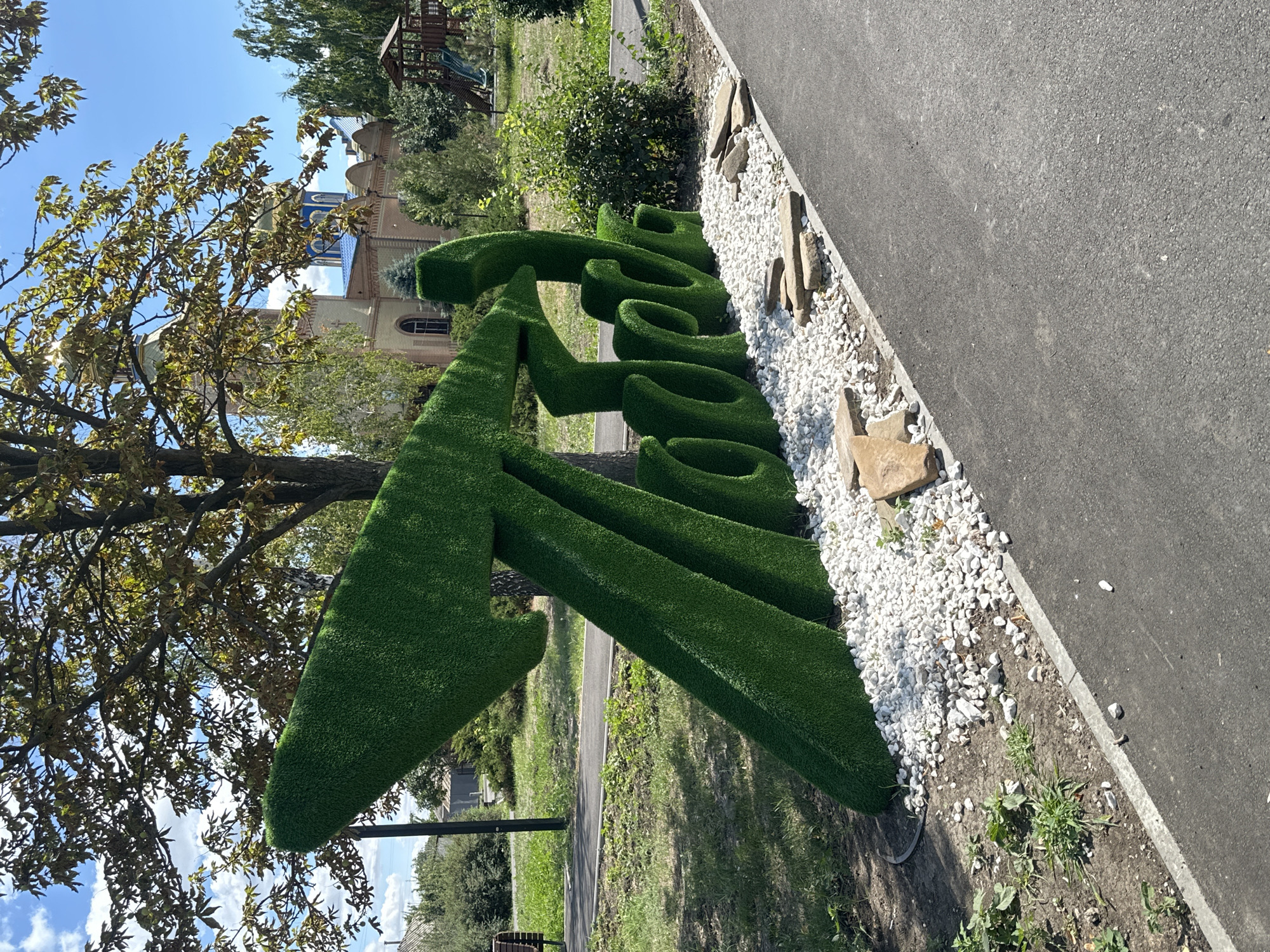 In Zhovte, Ukraine, a large, green, sculpted sign made of artificial turf stands in an outdoor park setting. The sign appears to be text, with curved and angular shapes forming letters. It is placed on a bed of white gravel and interspersed with flat, light-colored stones. Behind the sign, a paved path runs horizontally, and further back, a grassy area is visible with trees and a building with blue domes, likely a church. A children's playground structure with slides is also visible in the background. The sky is blue with scattered clouds, and the overall scene depicts a sunny day in a park.