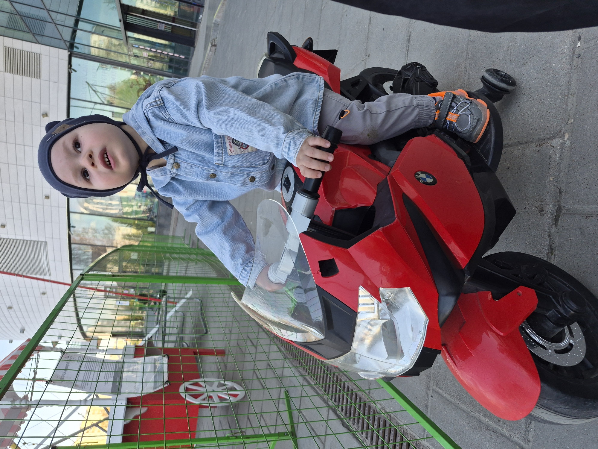A young child wearing a blue denim jacket and a dark blue hat is sitting on a red toy motorcycle. He has light-colored pants and gray and orange sneakers. The motorcycle is parked on a gray concrete surface. A green metal fence is visible on the left side of the image, and behind it, there is a building with glass windows. The scene is taken in Tobolsk, Russia.