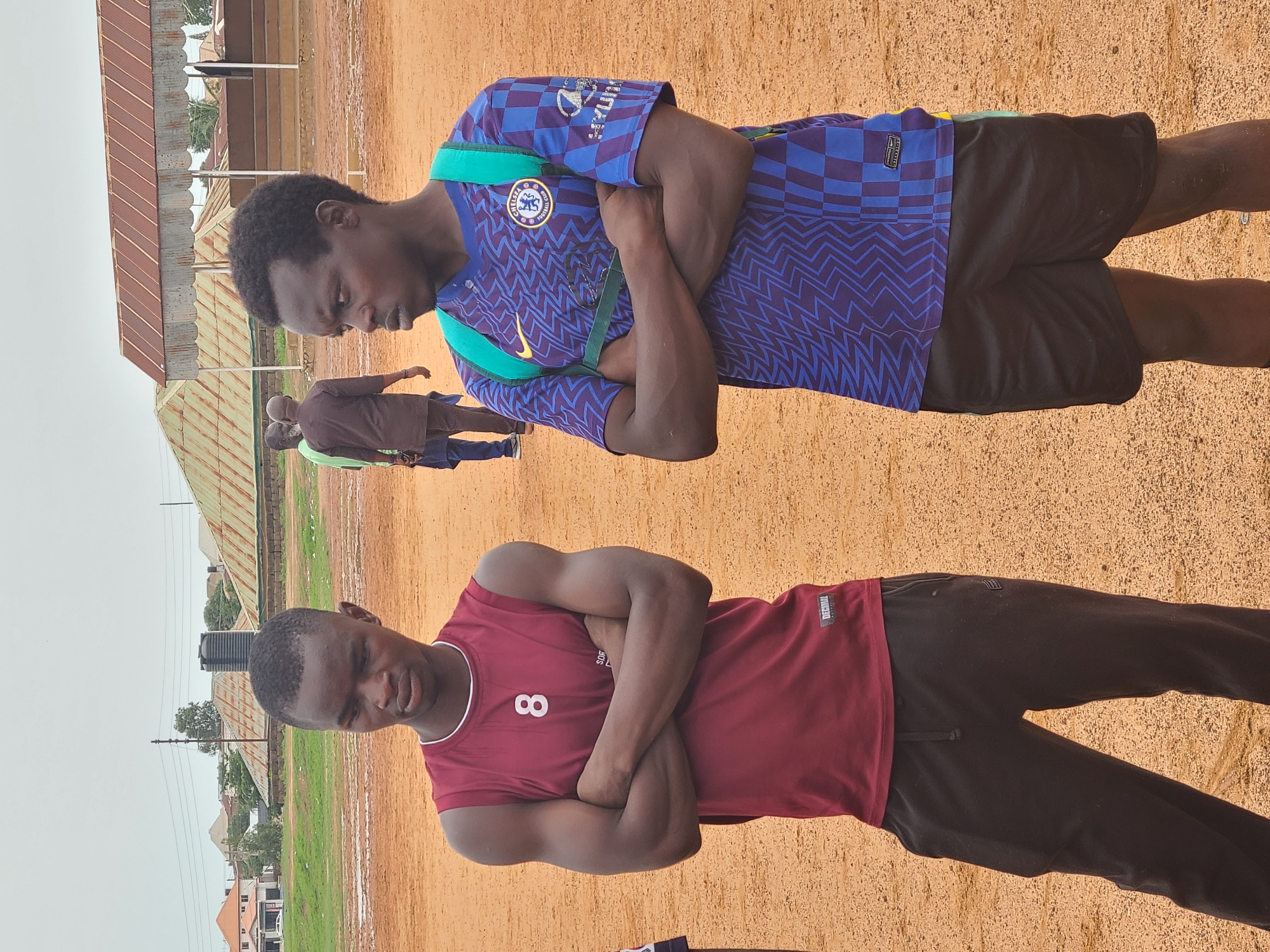 Two young men are standing outdoors on a bare, tan ground surface. One is wearing a purple and blue Chelsea soccer jersey with black shorts, and has his arms crossed. The other is wearing a maroon sleeveless shirt with the number 8 on it and dark pants, also with his arms crossed. In the background, there are buildings and other people. The location appears to be in the city of Unknown, Unknown.