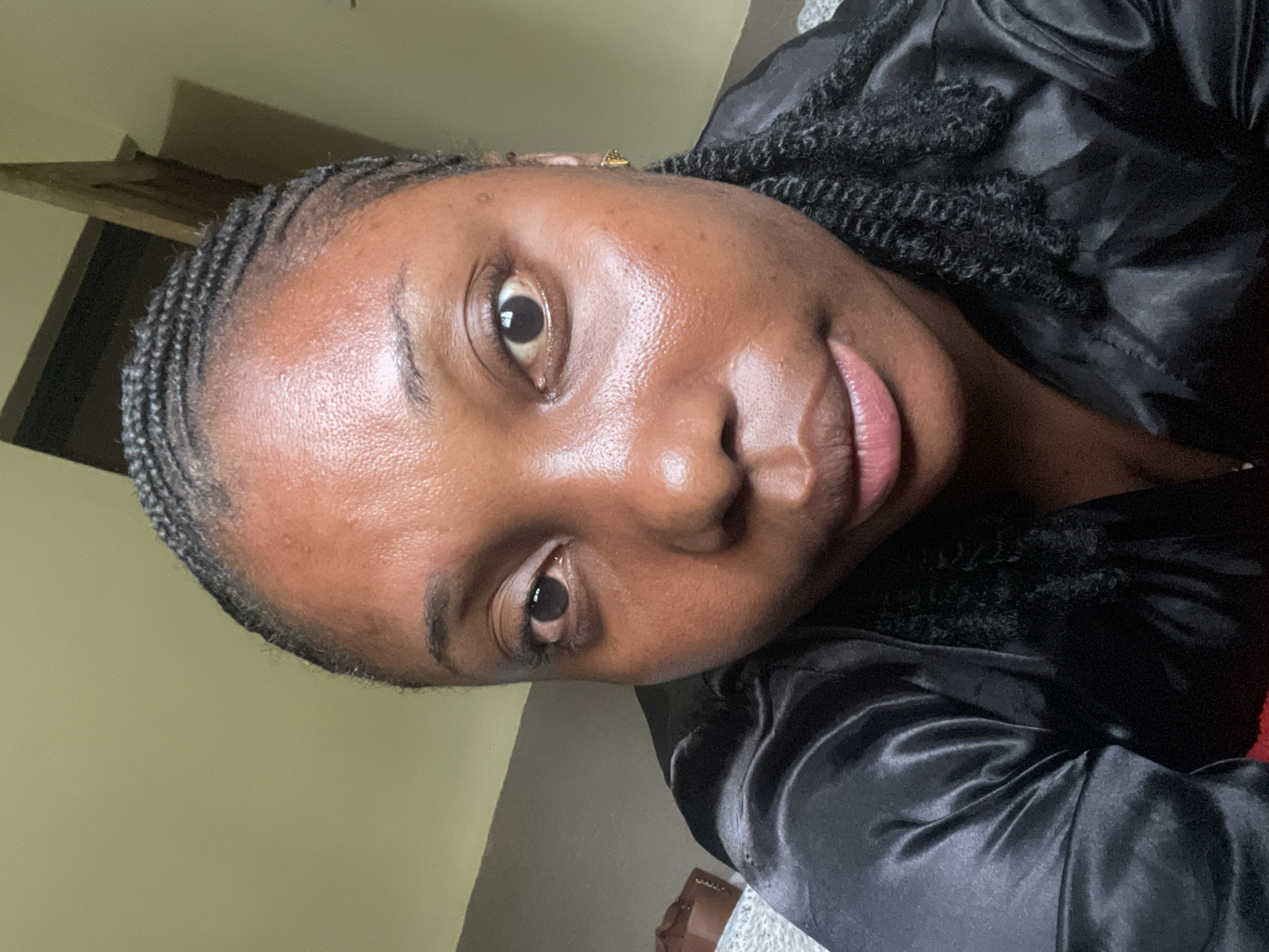 A rotated close-up portrait shows a young Black woman looking directly at the camera. Her dark skin has a natural sheen, particularly on her forehead and nose. Her dark eyes are open, and her lips are slightly parted, revealing a soft pink hue. Her hair is styled in cornrows at the front, transitioning into twisted or braided sections visible around her shoulders. She wears a black satin-like garment, possibly a robe or top. A small gold-colored earring is visible in her right ear. The background is simple and out of focus, appearing to be an indoor setting with plain walls and a hint of wooden furniture in the upper left. Her expression is calm and neutral. There is no visible text. The setting is indoors in what appears to be a relaxed environment.