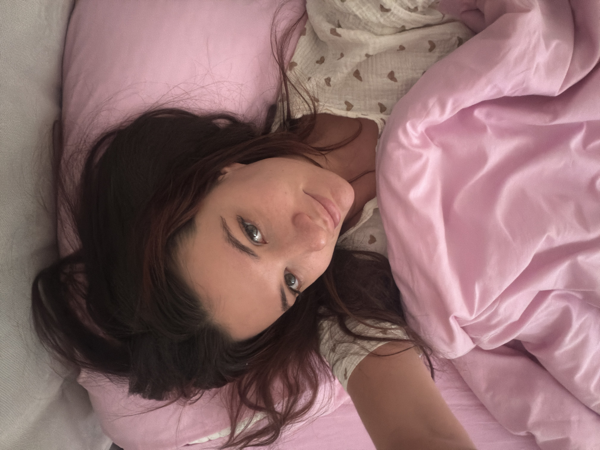 A young adult female is depicted lying supine in a bed, looking directly at the camera with a slight smile. Her long, dark brown hair is spread out on a light pink pillow. She has light-colored eyes and a fair complexion. She is wearing a light-colored top patterned with small heart shapes. A light pink duvet or blanket with a smooth, satin-like texture covers her lower body, displaying soft folds. The immediate background indicates an indoor setting, likely a bedroom, with a light-colored surface visible at the top edge of the frame. No visual information within the image provides details regarding its specific location within the city of Belgrade, Serbia.