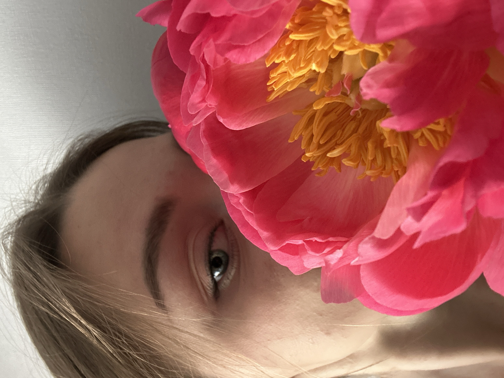 A close-up photograph depicts a fair-skinned person with light brown hair, partially obscured by a vibrant pink peony. The person's left eye is visible, appearing to be grey or blue. The peony flower is prominently featured, with layers of pink petals surrounding a cluster of yellow stamens. The background is out of focus and appears to be a light grey or white. The image is tilted to the side. There is no discernible context indicating a location in the city of Kazan, Russia.