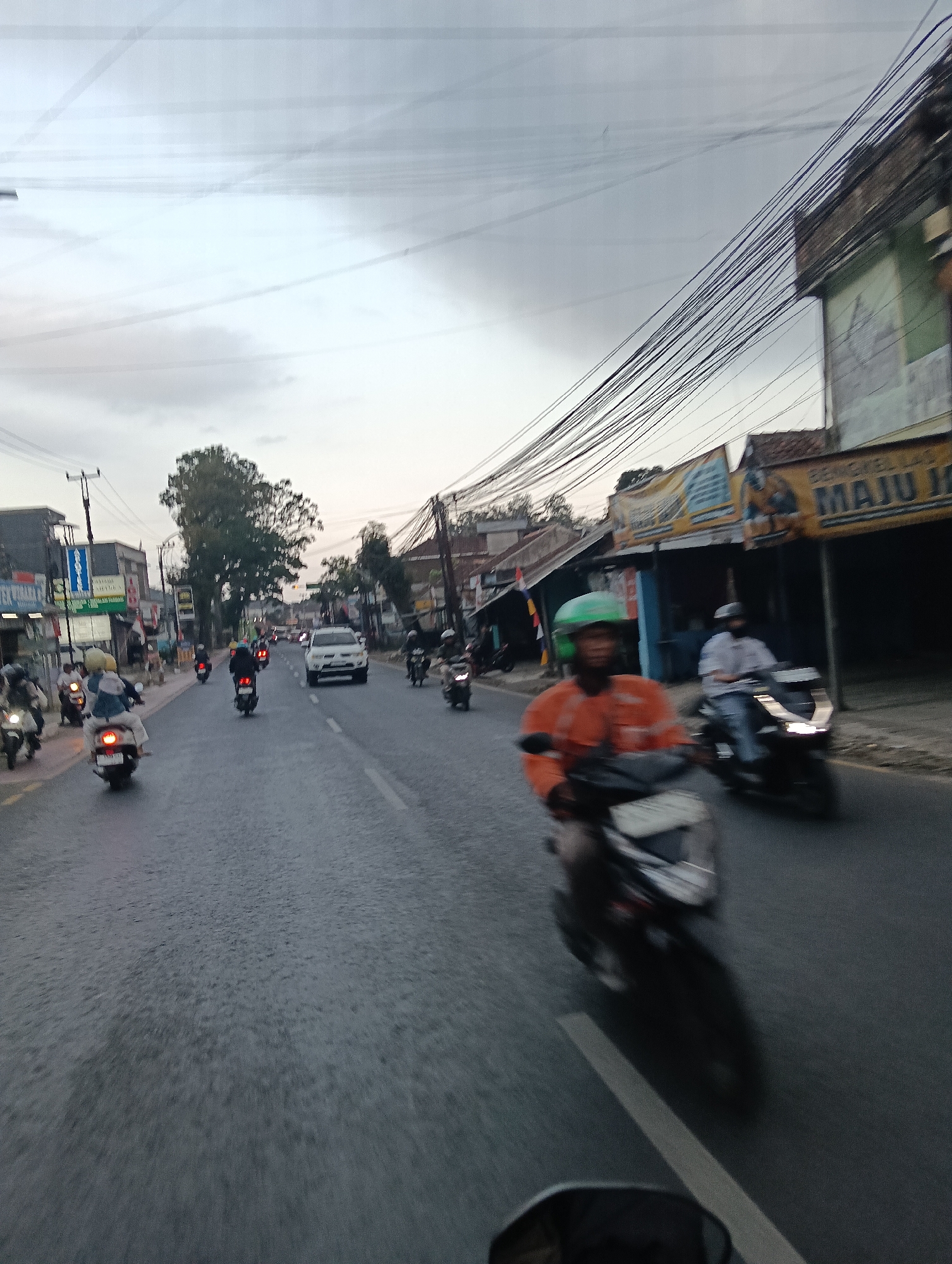 The image depicts a busy street scene in Tasikmalaya, Indonesia, likely in the late afternoon or early evening. The sky is overcast, with a gray hue. A significant number of motorcycles and a few cars are on the road, indicating moderate traffic. The buildings lining the street appear to be a mix of commercial establishments, with visible signage, including one with "MAJU JAYA" written on it. Numerous electrical wires are strung across the upper portion of the image, creating a cluttered overhead environment typical of urban Asian streets. A man in an orange jacket and green helmet is prominently featured on a motorcycle in the foreground, his face slightly blurred due to motion. Another person on a motorcycle is visible behind him, wearing a light-colored shirt. The road surface is asphalt and shows signs of wear. The overall impression is one of a typical bustling urban street in Indonesia.