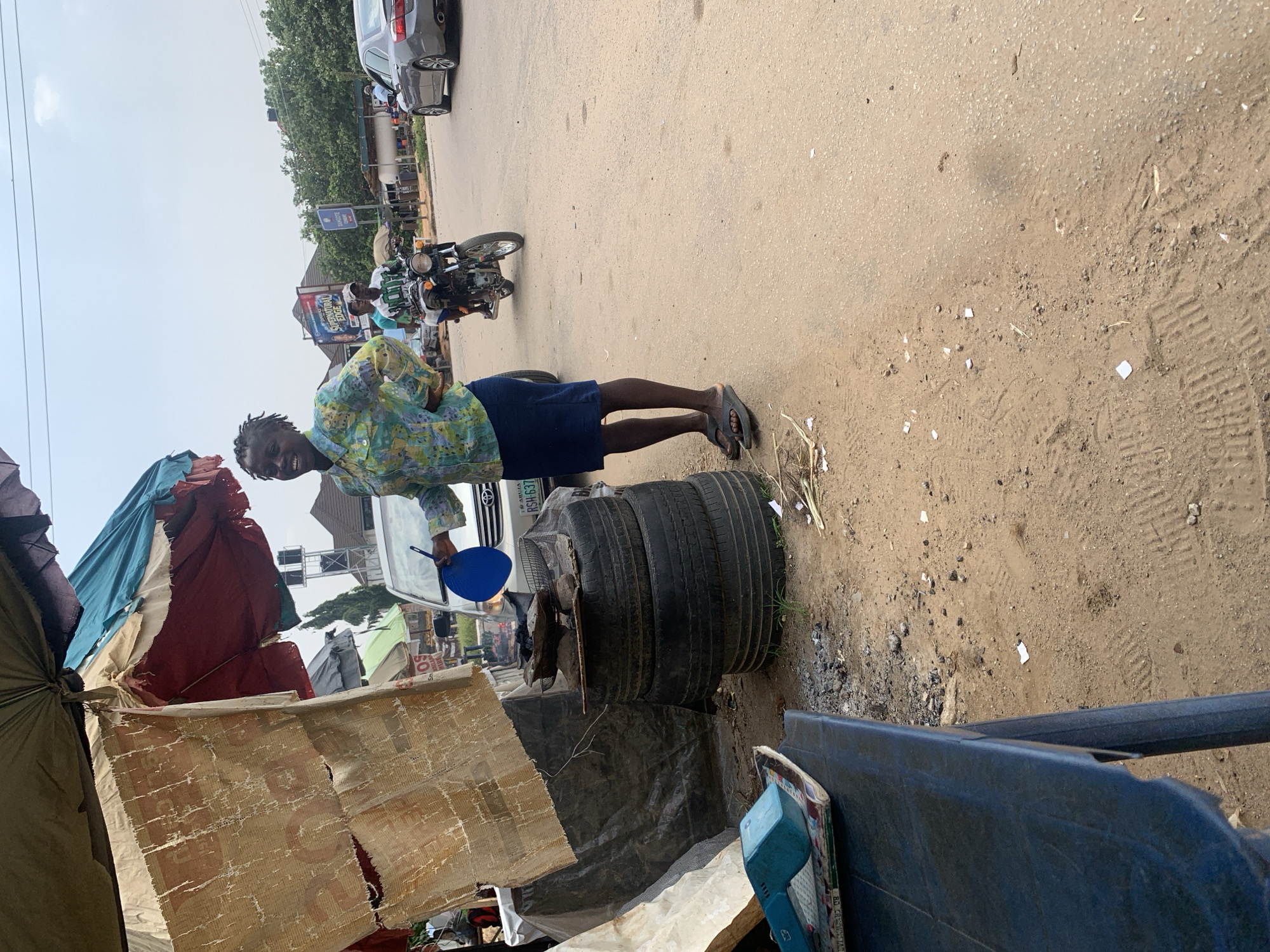 The image depicts an outdoor scene in Abuja, Nigeria. The foreground is dominated by various discarded materials, including stacks of tires, tarpaulin sheets, and what appears to be a damaged cardboard structure. A person, likely a male, is standing next to these items. He is wearing a colorful, patterned shirt and dark shorts, and black sandals. He is holding a blue fan in his right hand. Behind him, a white car with a Nigerian license plate (RSH-637) is visible. Further in the background, a motorcycle is parked, and other vehicles and buildings line a dusty road. Trees are visible in the distance, suggesting a partly urban, partly semi-rural setting. The overall impression is one of a street-level environment, possibly a market or a makeshift roadside stall, with signs of daily life and commerce. The lighting suggests it is daytime, with some sunlight casting shadows. The ground is predominantly dirt and asphalt, with scattered debris. There is no clear indication of the specific time of day or weather conditions beyond it being daylight. The mood is neutral, capturing a slice of everyday life in the city.
