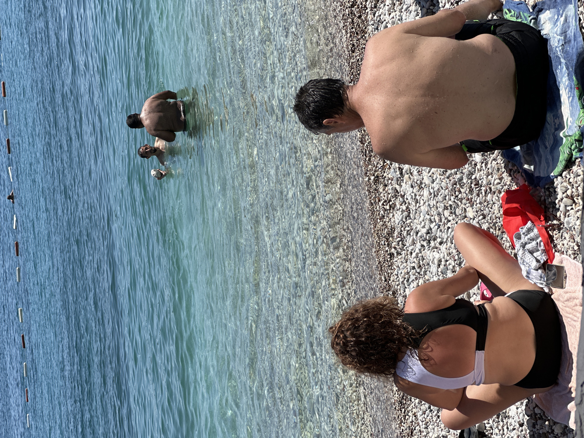 The image depicts a beach scene in Budva, Montenegro. In the foreground, a woman with curly brown hair is seated on a pebble beach, wearing a black and white bikini. Beside her, a man is also seated, facing away from the camera, wearing dark shorts. In the mid-ground, two men are visible in the clear turquoise water. One man, holding a light-colored object, is facing another man who is partially submerged. In the background, a line of buoys stretches across the water. The sea is calm, with sunlight glinting off the surface.