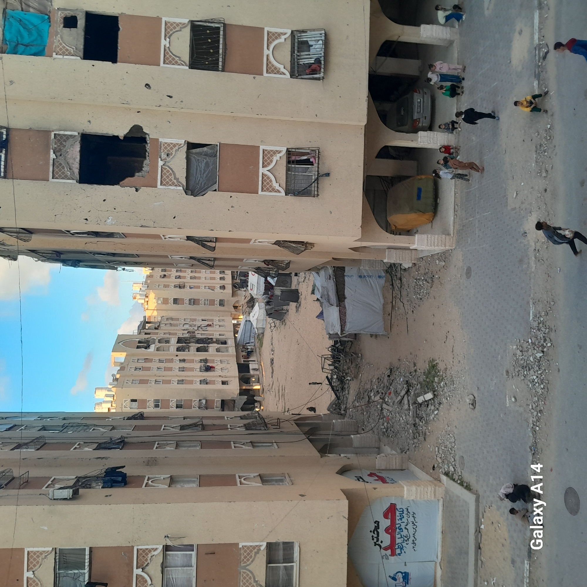 The image depicts a damaged urban environment, likely a residential area, with multi-story apartment buildings flanking a street. The scene is viewed from an elevated perspective, looking down.
The primary subjects are the numerous light-colored apartment buildings. Many windows are visibly broken, boarded up, or show dark, empty interiors, indicating damage. Some windows retain decorative frames, while others have metal grates. Clothes and other items hang from some upper windows. Below the buildings, the ground is a mix of paved street, which appears worn and scattered with small debris, and a wider dirt or gravel area. This area is extensively littered with rubble and larger pieces of construction waste.
In the rubble-strewn open space between the buildings, several temporary tent-like structures are set up, some of which prominently display the "unicef" logo, suggesting humanitarian aid or makeshift housing. A light-colored car is parked under an arched entrance of one of the buildings.
Several people, including adults and children, are present on the street. They are seen walking, standing, and gathering in small groups. One man in a dark shirt is walking away in the foreground, and other individuals and children are scattered further down the street, seemingly going about daily activities despite the damaged surroundings.
The time of day appears to be daytime, with a clear blue sky and some white clouds visible, providing natural light to the scene. The overall impression is one of a community enduring and adapting amidst significant destruction.
Visible text includes a "Galaxy A14" watermark in the bottom right corner of the image. On a large banner attached to one of the buildings, Arabic text is present, translating to: "Laboratory Hamad for all medical analyses. Address: Hamad City, Tower 5. Working hours: from 8 AM to 2 PM." The "unicef" logo is also visible on some of the temporary structures.