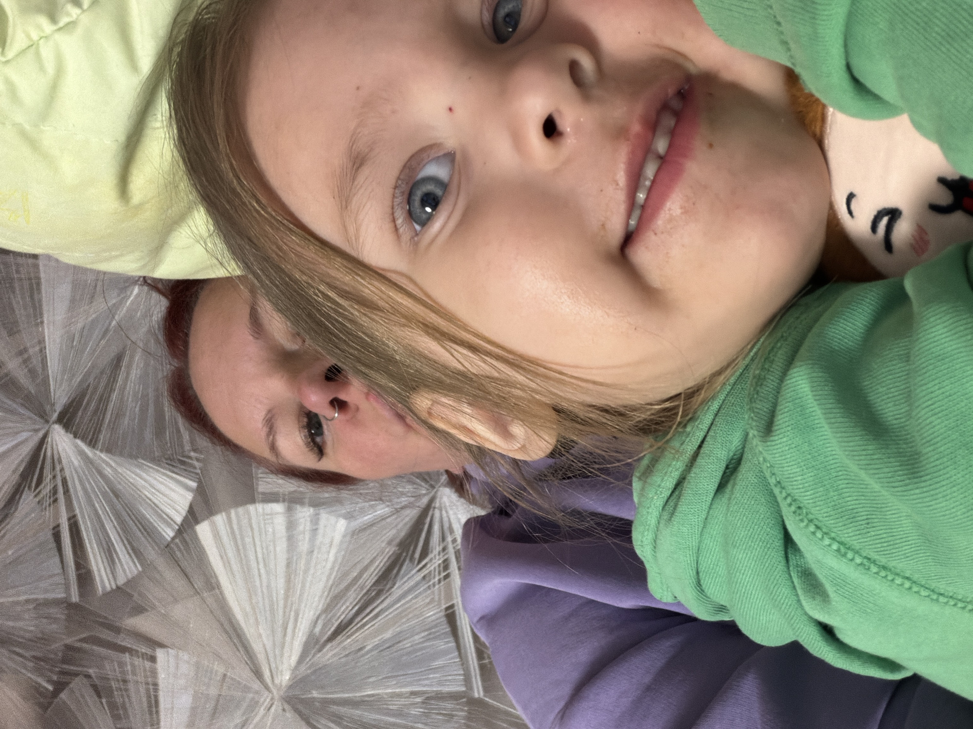 A close-up shot of a fair-skinned, blue-eyed child lying on a bed or floor with an adult. The child is wearing a green hooded sweatshirt and holding a stuffed animal toy. The adult, with reddish-brown hair and a nose ring, is lying behind the child on a patterned, white and gray sheet or blanket. The lighting is natural, and the photo appears to be taken indoors. There is a green pillow near the top left of the image. There is no explicit information to confirm the location is Chaikovsky, Russia, so this is not included in the description.