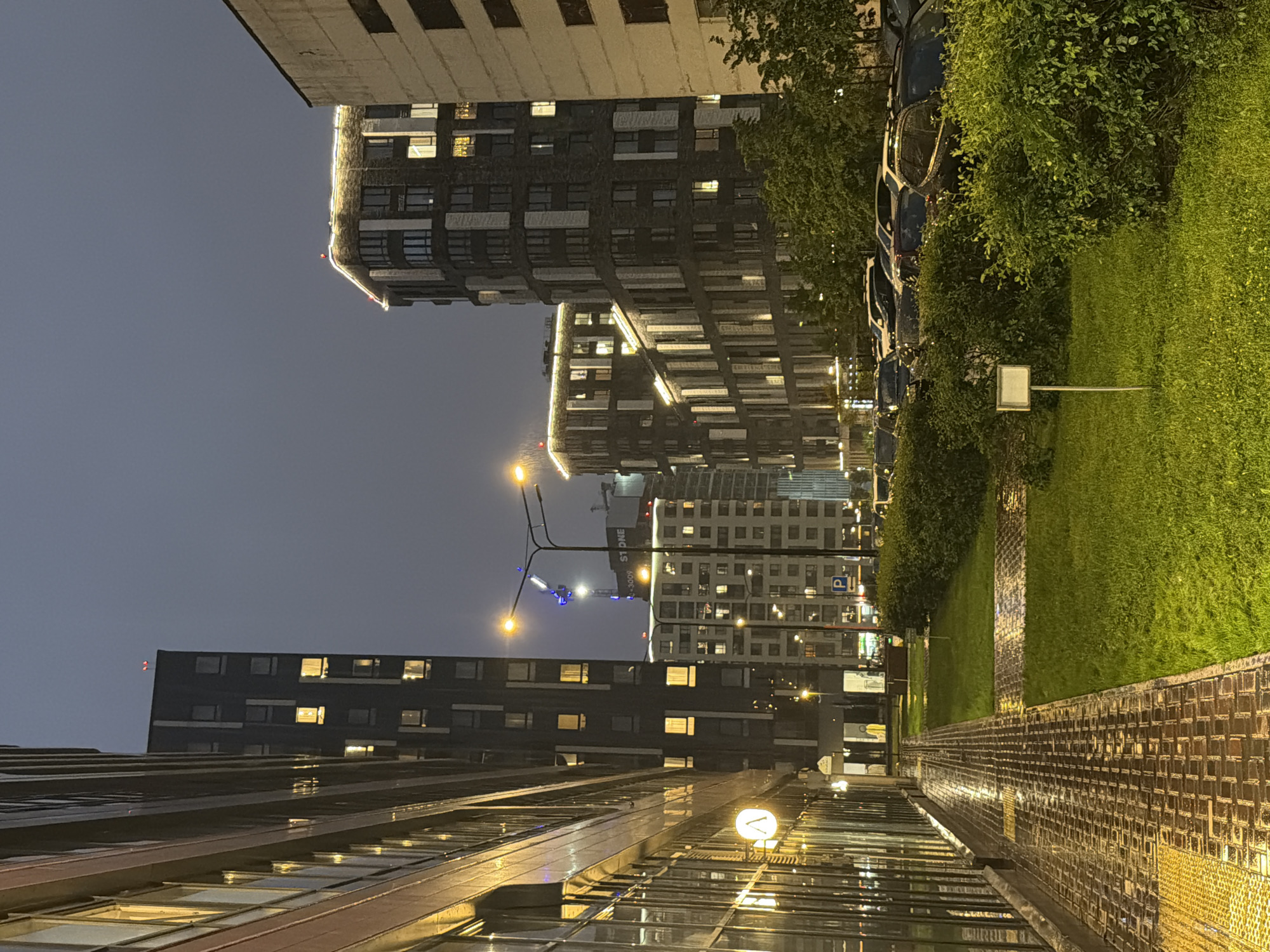 The image depicts a nighttime scene in a modern urban environment, likely Moscow, Russia, characterized by contemporary architecture and lush greenery. The main subjects are several tall, multi-story buildings with numerous illuminated windows, suggesting residential or office spaces. These buildings feature dark facades with contrasting white or light-colored horizontal bands and decorative lighting along their upper edges.
To the right of the buildings is a well-maintained green lawn, bordered by a brick pathway that appears wet, indicating recent rain or damp conditions. A row of dark-colored cars is parked alongside a dense line of trees and bushes separating the parking area from the buildings. Streetlights with bright yellow bulbs illuminate parts of the scene, casting a warm glow on the wet surfaces.
The time of day is clearly night, evidenced by the darkness of the sky and the artificial lighting. The weather appears to be humid or wet, as suggested by the sheen on the pavement and the reflections on the cars. The overall mood is quiet and perhaps a little melancholic, with the stark architectural lines softened by the warm light and the natural elements.
Visible text includes a "P" symbol on a blue square sign, indicating parking, and what appears to be a partial sign on one of the buildings that reads "S1 ONE" or similar. The reflective surfaces of the buildings' windows and the wet ground create interesting patterns of light and shadow. The image is shot from a low angle, emphasizing the height of the buildings and the expanse of the sky.