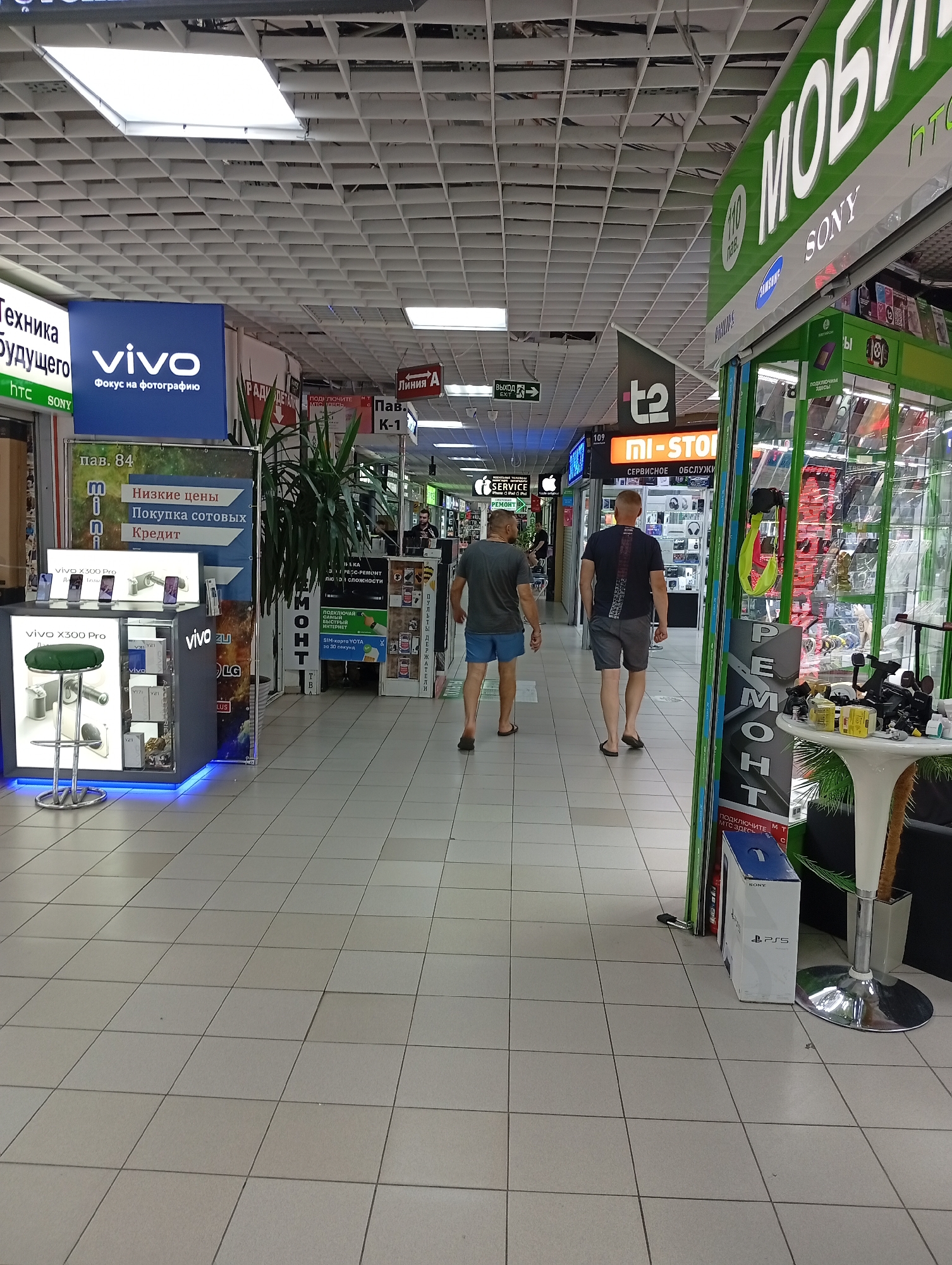 The image shows an indoor market or shopping center. A man in a dark t-shirt and blue shorts walks away from the camera towards the right side of the image, with another man in a black t-shirt and grey shorts walking ahead of him. In the background, there are several shops with signs in Russian, including one that says "VIVO" with "Focus on photography" below it, and another that says "MI-STORE". There is also a display with phones and accessories for sale. To the right, there is a stall with various electronic items and a white table with cameras on it. A sign that says "РЕМОНТ" (repair) is visible. A white box with "PlayStation 5" is on the floor. The environment is lit by overhead fluorescent lights, and the floor is tiled.