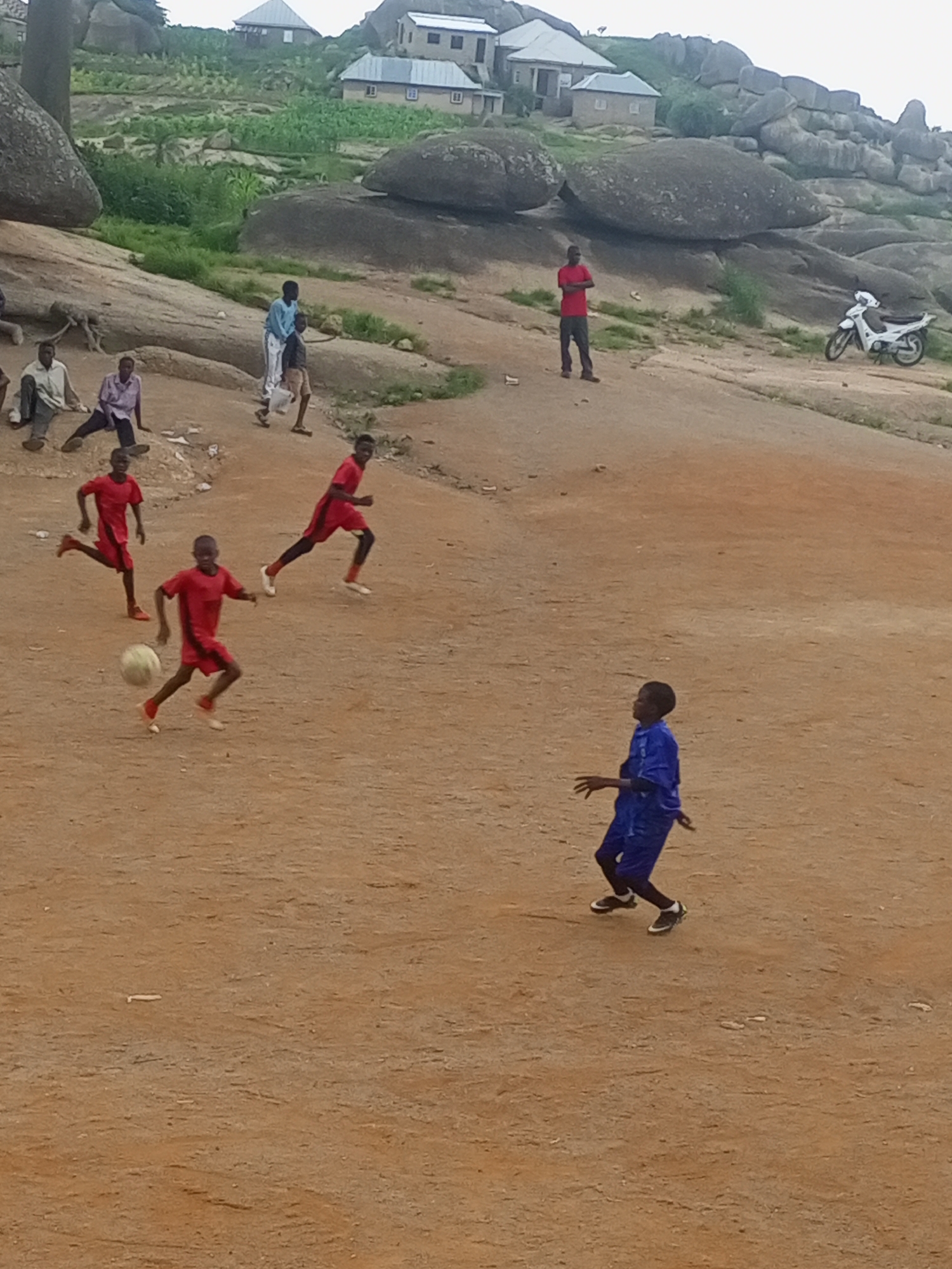 The image depicts a scene in Jos, Nigeria, likely on a rocky terrain with sparse vegetation. In the foreground, several young boys are engaged in a game of soccer on a dusty, reddish-brown field. Three boys in red jerseys and shorts are actively playing, with one boy in the center dribbling a ball. Another boy in a blue jersey and shorts stands to the right, appearing to be preparing to receive or interact with the ball. In the background, there are other individuals observing the game or resting, some seated on the rocky ground. Further in the distance, houses are visible on a hillside, characteristic of Jos's landscape, with large rock formations dominating the upper parts of the scene. A white motorcycle is parked on the rocky ground in the middle ground, suggesting the presence of transportation. The sky is overcast, indicating a possibly hazy or cloudy day.