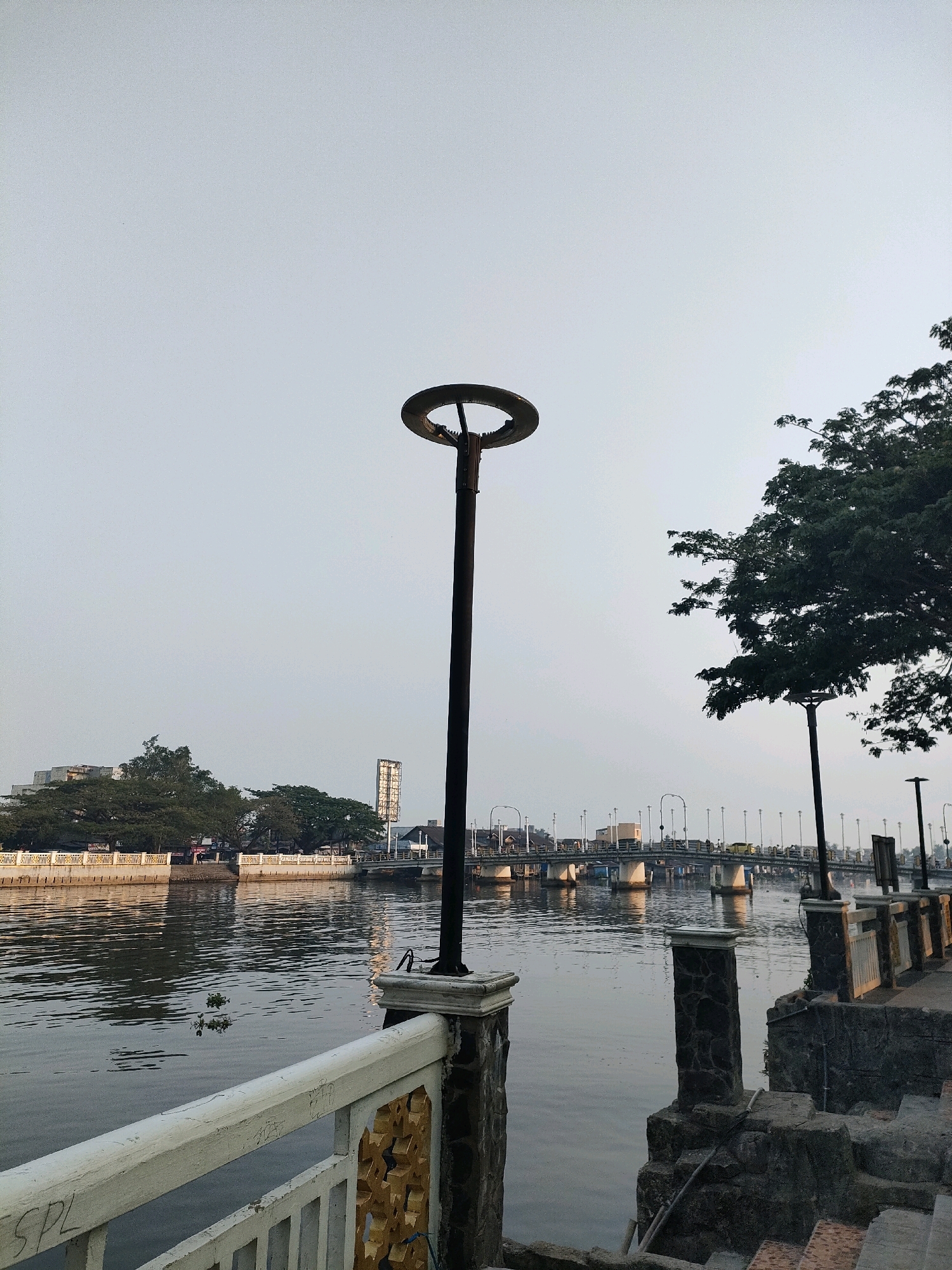 This image depicts a waterfront scene in Banjarmasin, Indonesia, likely along the Martapura River. In the foreground, a white railing with gold decorative elements runs along a stone embankment. A tall, dark lamppost with a circular, open top stands prominently on the embankment, supported by a stone pillar. To the right, a concrete pathway leads down a set of stairs.
Across the river, the opposite bank is lined with trees and buildings, including a tall, rectangular structure with a billboard-like display. A bridge spans the river in the middle ground, with traffic and streetlights visible on it, suggesting it is a busy area. The water's surface is slightly rippled, reflecting the overcast sky. The overall atmosphere is calm and subdued, possibly indicating early morning or late afternoon light. No people are clearly discernible in the image.