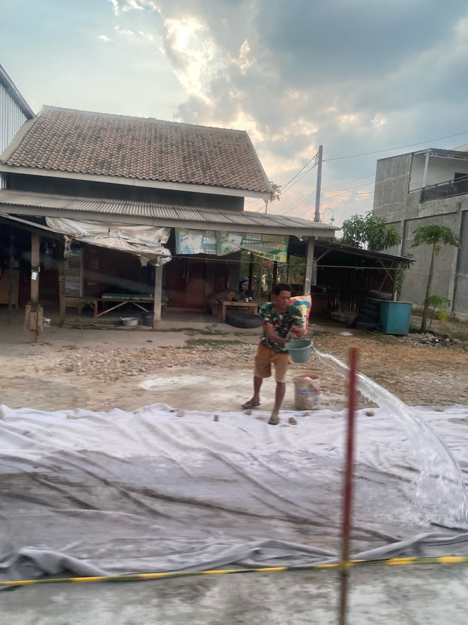 A man in a camouflage t-shirt and tan shorts is actively pouring water from a green bucket onto a white tarp that is spread out on the ground. He is standing on a dirt and gravel surface in front of a collection of buildings. The buildings appear to be a small shop with a corrugated metal roof and a two-story concrete structure in the background. A large number of stacked tires are visible behind the shop, as well as a blue plastic drum. The sky is cloudy and appears to be late in the day. The overall scene suggests an outdoor working area or a street in a developing area, possibly in Banjar Sari, Indonesia.