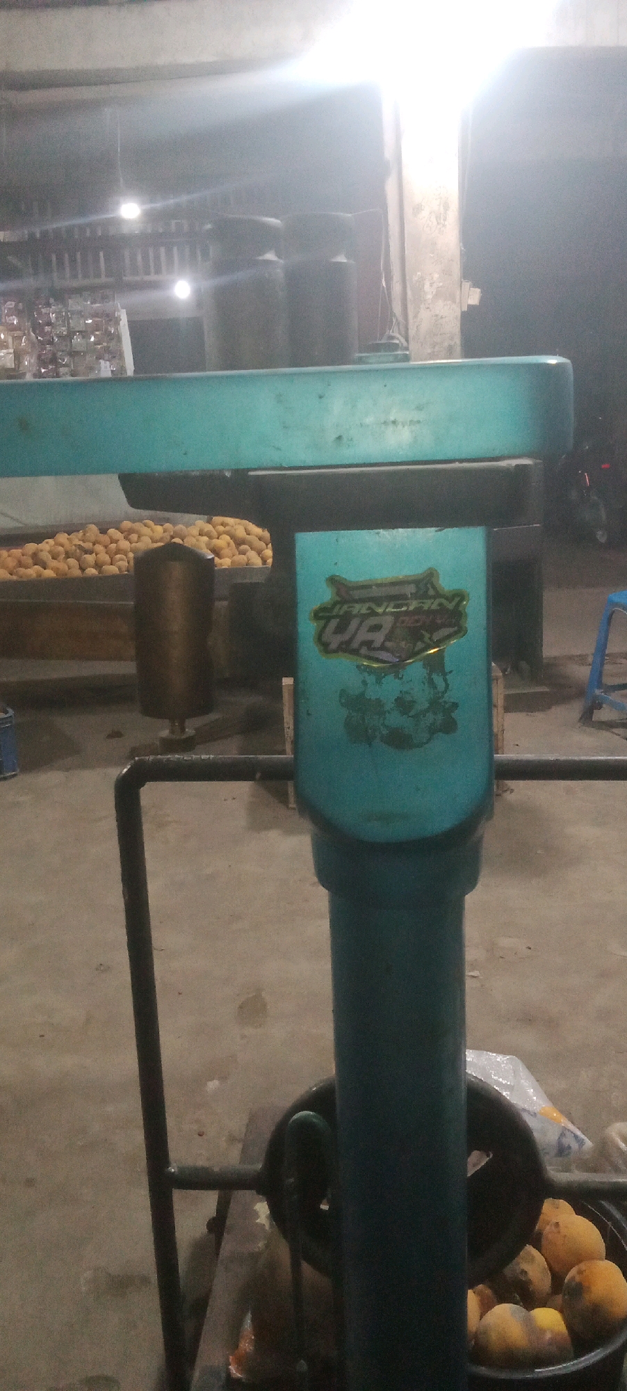 This is a vertical, medium-shot, dimly lit photograph taken at ground level, likely in Karawang, Indonesia, depicting an outdoor or semi-outdoor setting with a focus on a teal-colored machine and its surroundings. The machine, which appears to be some sort of processing or dispensing equipment, has a prominent teal-colored horizontal bar on top and a vertical cylindrical component in the foreground. This vertical component has a sticker with a graphic and text on it, appearing to be in a language that could be Indonesian.
To the left of the main machine, a large bin filled with what appear to be yellow fruits, possibly potatoes or mangoes, is visible. Another metal cylindrical object, likely part of another machine, is situated behind the fruit bin. In the background, the scene is blurry, but suggests shelves with items, possibly merchandise, and several large, dark cylindrical containers. Bright lights are visible in the upper background, illuminating the scene. A motorcycle is parked in the far right background, and a blue stool is also present. The ground is a concrete surface, showing some dirt and debris. The overall atmosphere is utilitarian and appears to be a commercial area, possibly a market or roadside stall.