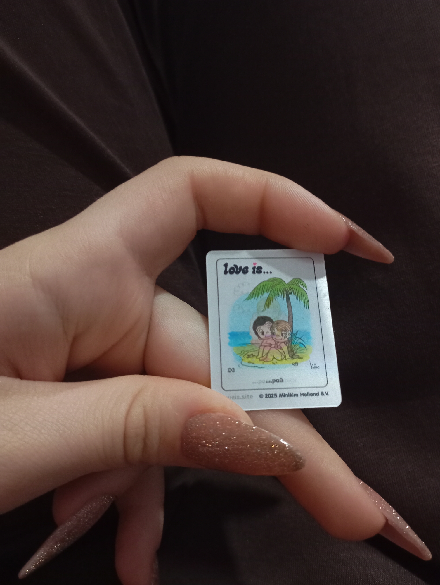 A hand holds a small "Love is..." card featuring a cartoon illustration of a couple under a palm tree on a beach. The card includes copyright information "© 2025 Minikim Holland B.V.". The background is out of focus, but appears to be a dark brown fabric. The nails of the hand are long and glittery brown. There is no indication of the location being Krasnodar, Russia.