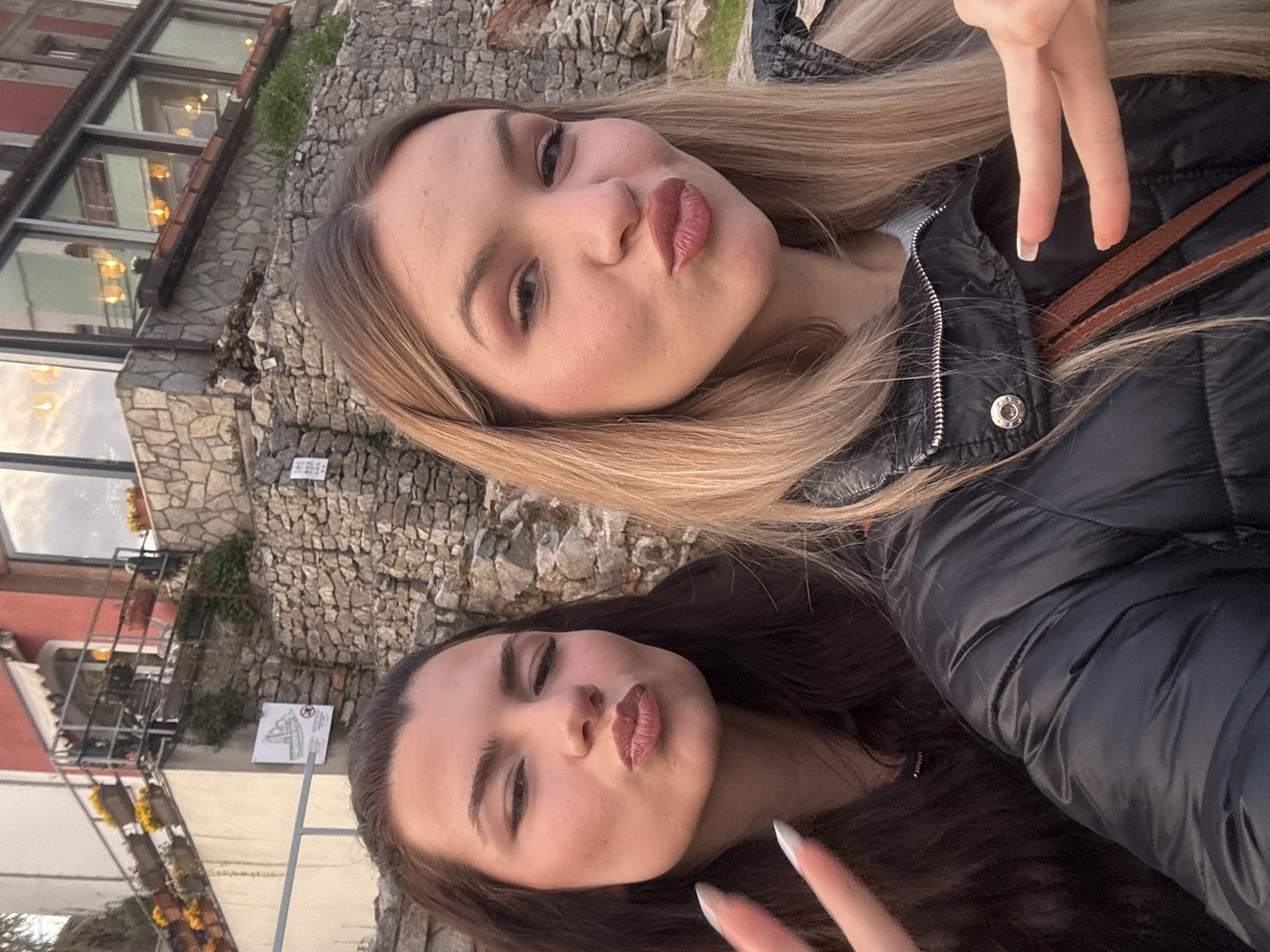 Two young women are taking a selfie in what appears to be the Old Town of Plovdiv, Bulgaria. The woman on the top has blonde hair and is wearing a black puffer jacket. The woman on the bottom has dark hair and is also wearing a black puffer jacket. Both are making a "duck face" pose. Behind them is a stone wall and buildings with windows, indicative of the architectural style of the Old Town. There is a sign on a white background visible in the background. The weather appears mild.