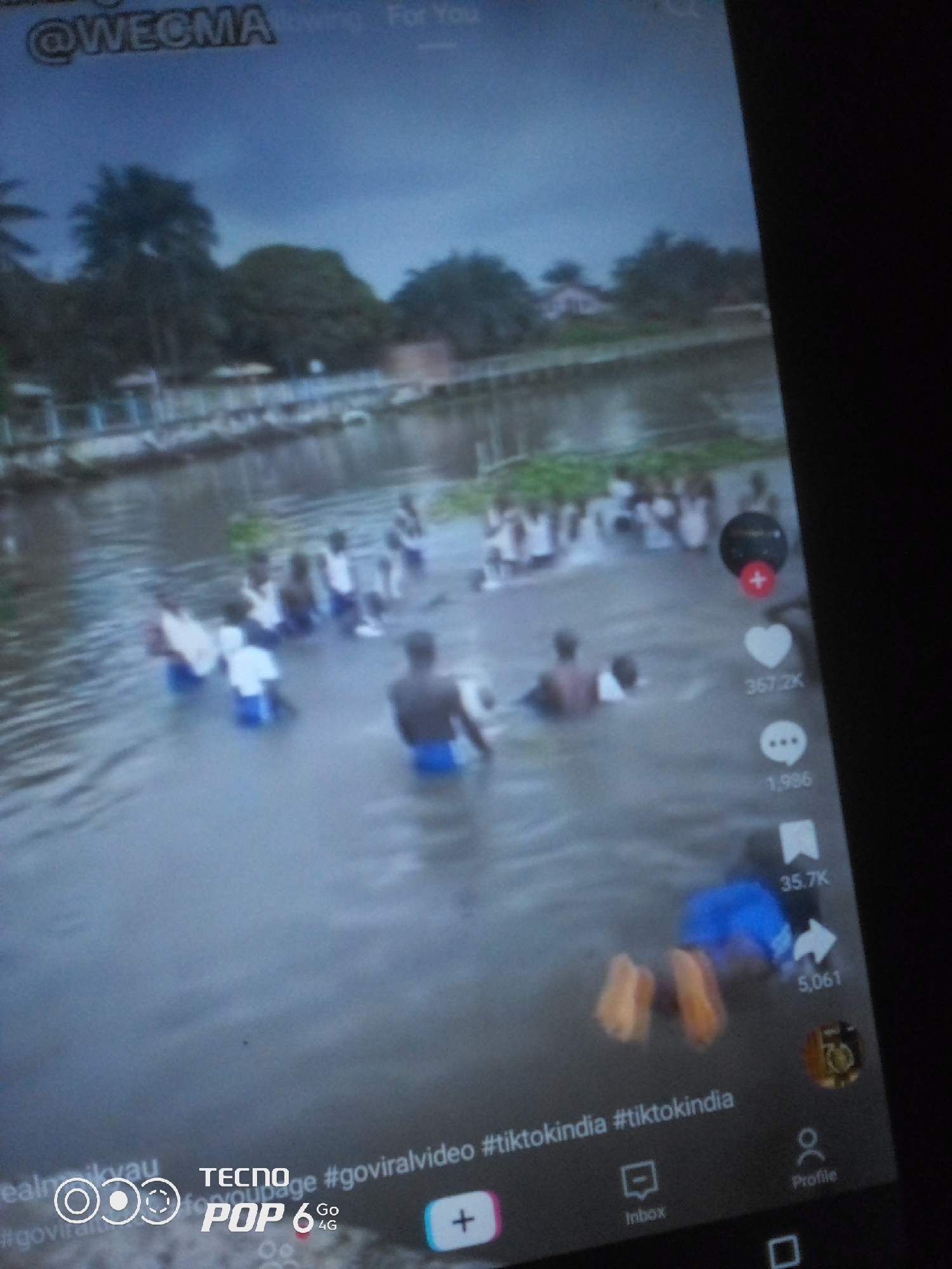 A group of people are wading in shallow water. The scene appears to be an outdoor location with trees and a building in the background. The water is murky and the ground beneath appears to be dirt or mud. The people are dressed in casual clothing, with some wearing t-shirts and shorts, and others wearing what appear to be blue bottoms. Some are standing, while others are sitting or crouching in the water. The overall atmosphere suggests a casual gathering or activity.