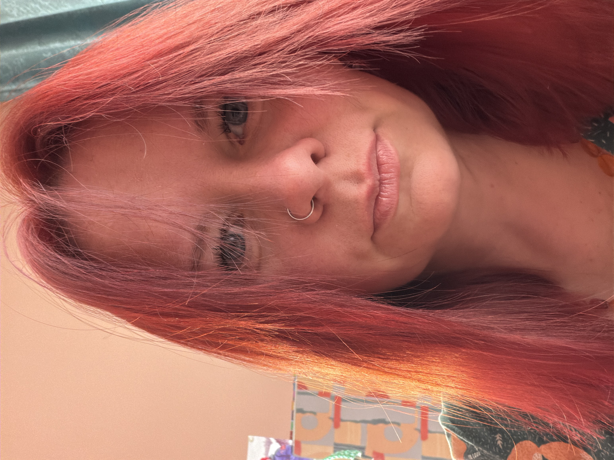 A close-up shot of a woman with bright pink hair, wearing a nose ring. The background is blurred, but there are hints of a teal-colored curtain and a patterned object. The woman's face is slightly angled towards the viewer, and she has a subtle smile. The lighting is soft and diffused. It is not possible to determine the location based on the image provided.