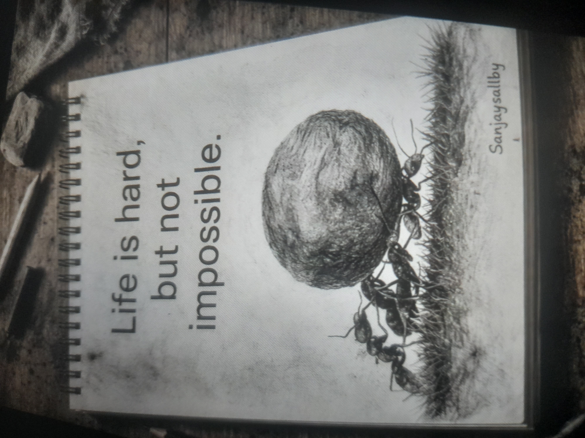 A close-up, slightly angled photograph captures a notebook displaying a motivational image. The notebook, bound by a metal spiral on the left, features the text "Life is hard, but not impossible." The image accompanying the text depicts several ants working together to push a large, round object, resembling a boulder, up a grassy hill. "Sanjaysallby" is written vertically on the right side of the page, likely indicating the artist's name. An eraser and pencil rest on the wooden surface next to the notebook, suggesting an artistic or creative workspace. The overall tone of the image is monochromatic, with shades of gray dominating the composition. The location in Akwanga, Nigeria is not visible in the image.