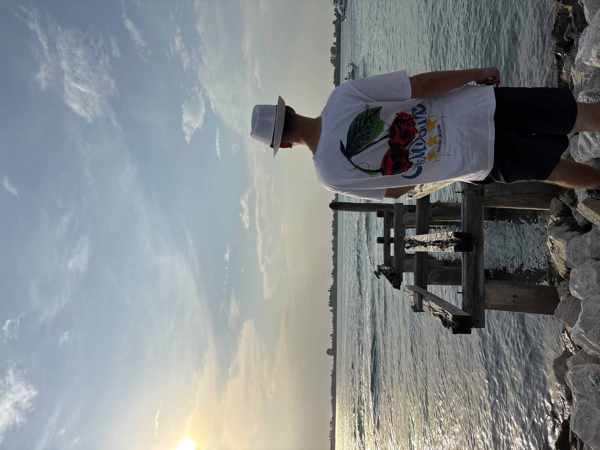 A person wearing a white shirt, black shorts, and a white hat stands on a wooden pier in Punta Sabbioni, Italy, looking out over the water. The sky is pale blue with scattered clouds, and the sun is low on the horizon. The water reflects the sunlight, creating a shimmering effect. The pier is made of weathered wood and appears old. Rocks line the edge of the water near the pier. The person is facing away from the camera. In the distance, the shoreline and a few boats are visible.