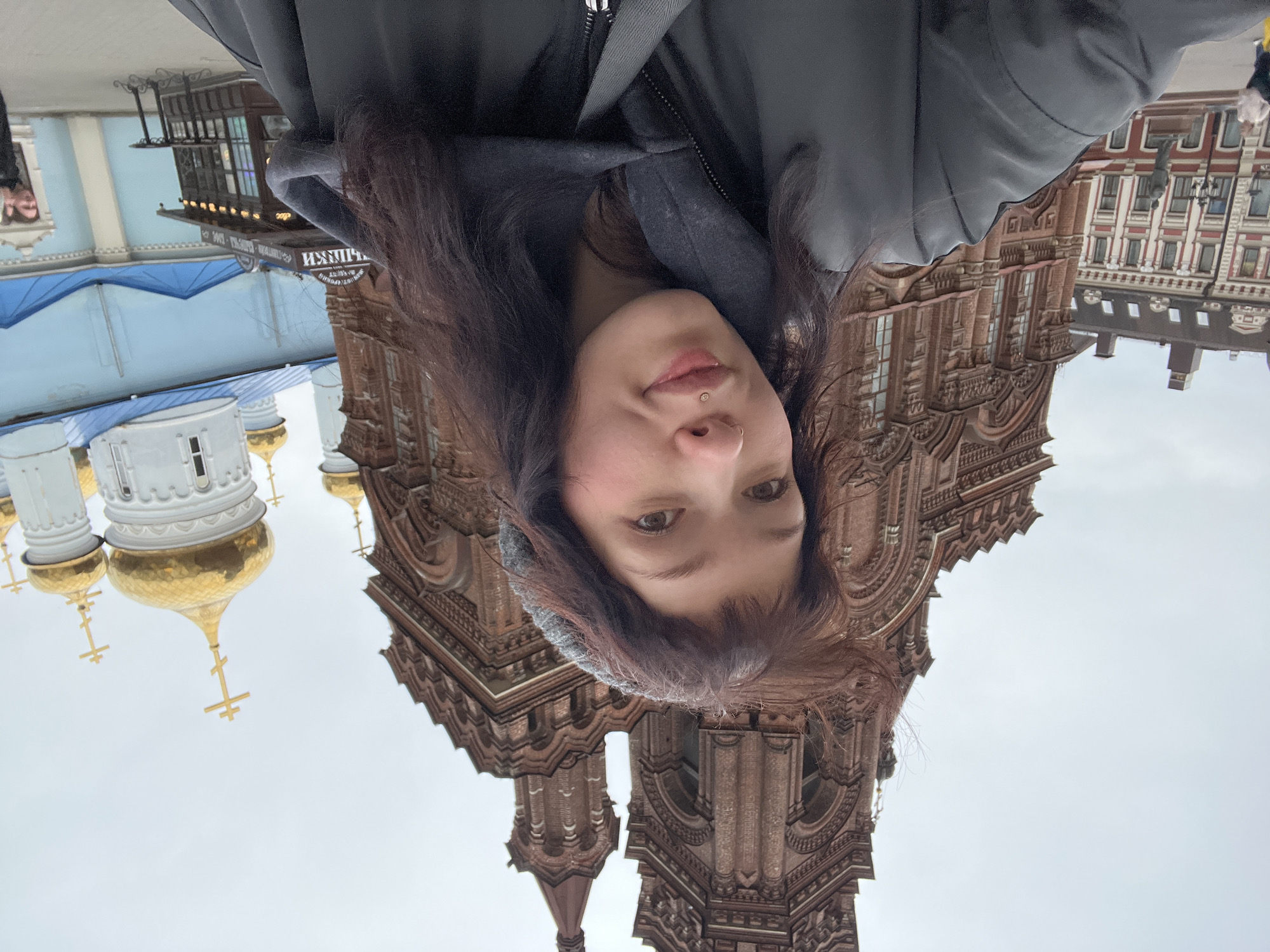 The image is an upside-down selfie of a woman with long brown hair, a nose piercing, and a lip piercing. She's wearing a dark jacket. The background is a cityscape in Kazan, Russia. Distinctly visible is the Kul Sharif Mosque with its blue roof, white walls, and golden domes. Also in the background is the Annunciation Cathedral with brick architecture and a building featuring red and white facade elements. The sky is overcast.