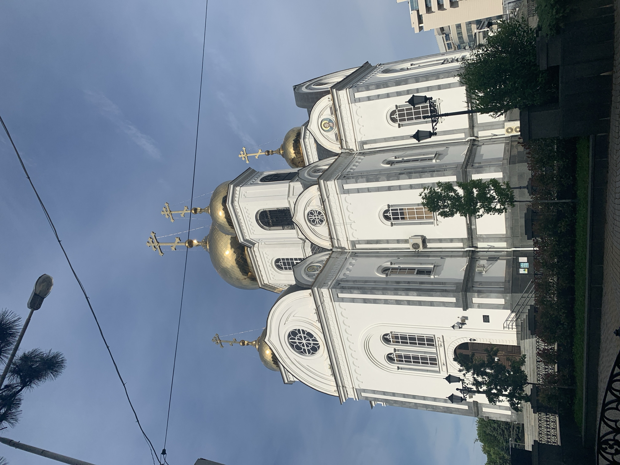 The image shows a tall, white Orthodox church with golden domes in the city of Unknown, Unknown. The church has multiple levels, each decorated with arched windows and intricate detailing. The golden domes are topped with Orthodox crosses. In the foreground, there are trees and a lamp post. The sky is blue with some wispy clouds. An air conditioning unit is visible on the exterior of the church. The church is located in an urban area with a building visible in the background.